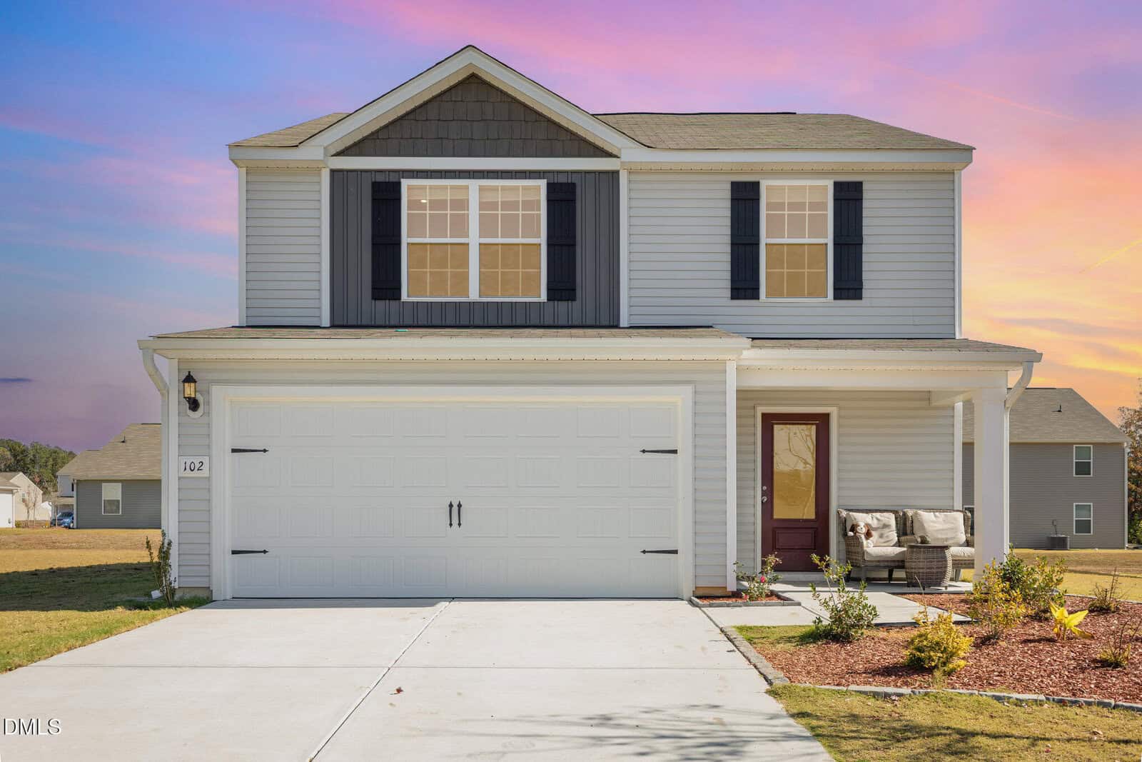 A two-story Oxford suburban house with gray siding, black shutters, and a double garage rests at 102 Cultivar Street. A covered porch with seating sits on the right. The sunset sky accents the landscaped yard of grass and small plants.