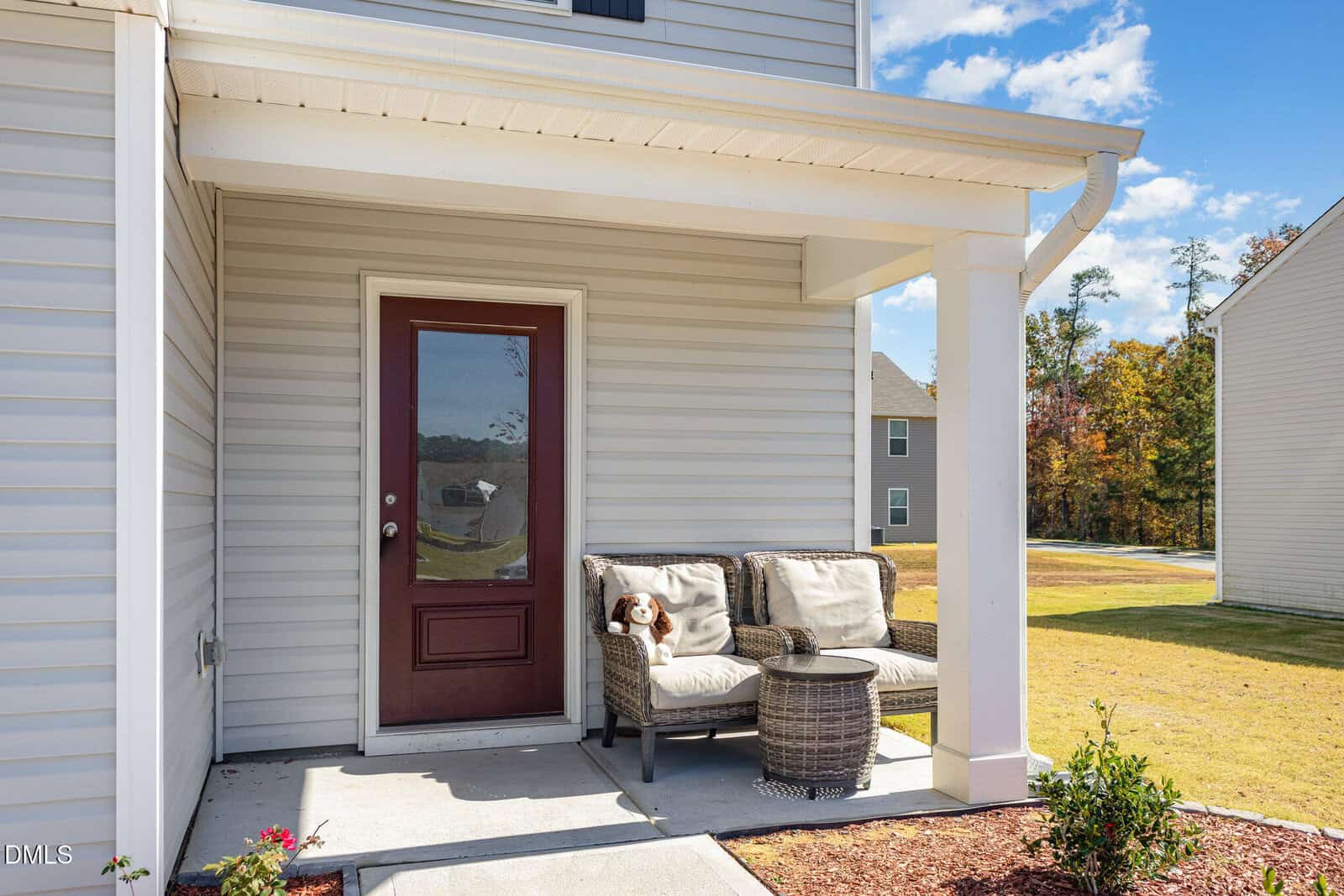 A small covered porch with a red door, two cushioned chairs, a round wicker table, and a stuffed animal on one chair. The house exterior is light-colored siding, and a grassy yard is visible.