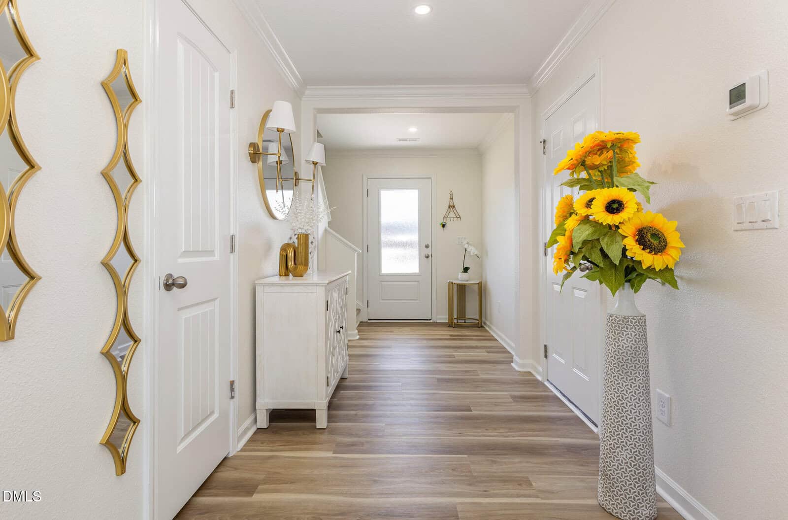 A bright hallway with light wood flooring, cream walls, a white front door, a sideboard with a round mirror and wall lamps, a decorative gold wall piece, and a tall vase of sunflowers.