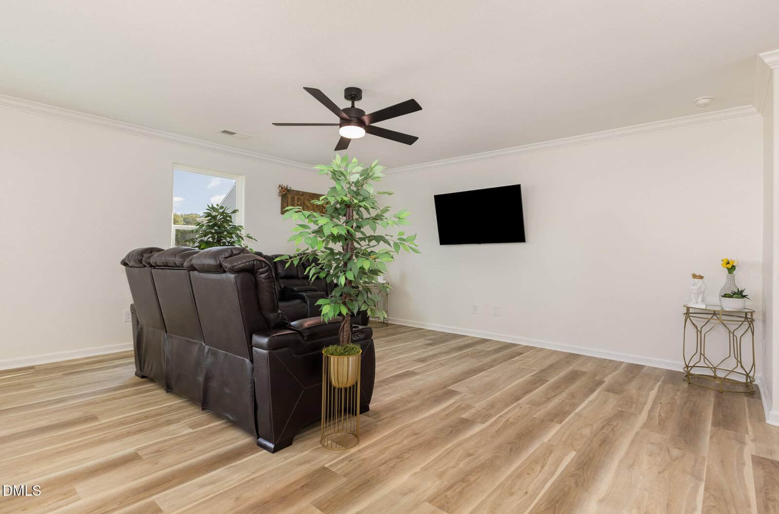 A living room with light wood flooring, a dark brown leather sofa, a ceiling fan, a wall-mounted TV, a potted plant, a small side table with decor, and a window letting in natural light.