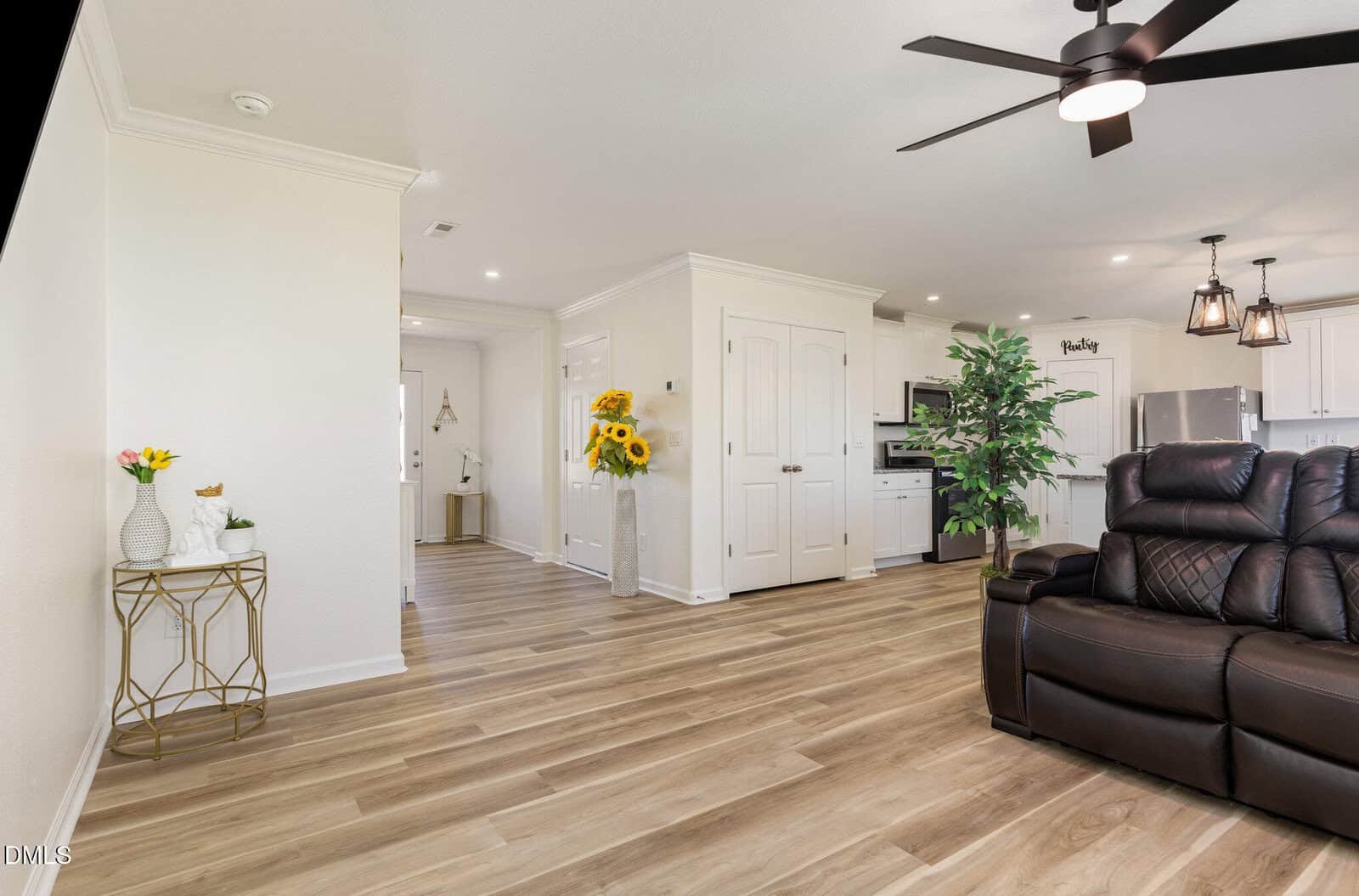 A modern living area with light wood flooring, white walls, a dark brown recliner, a gold accent table with decor, artificial plants, and a view into a white kitchen with pendant lights and a double-door pantry.