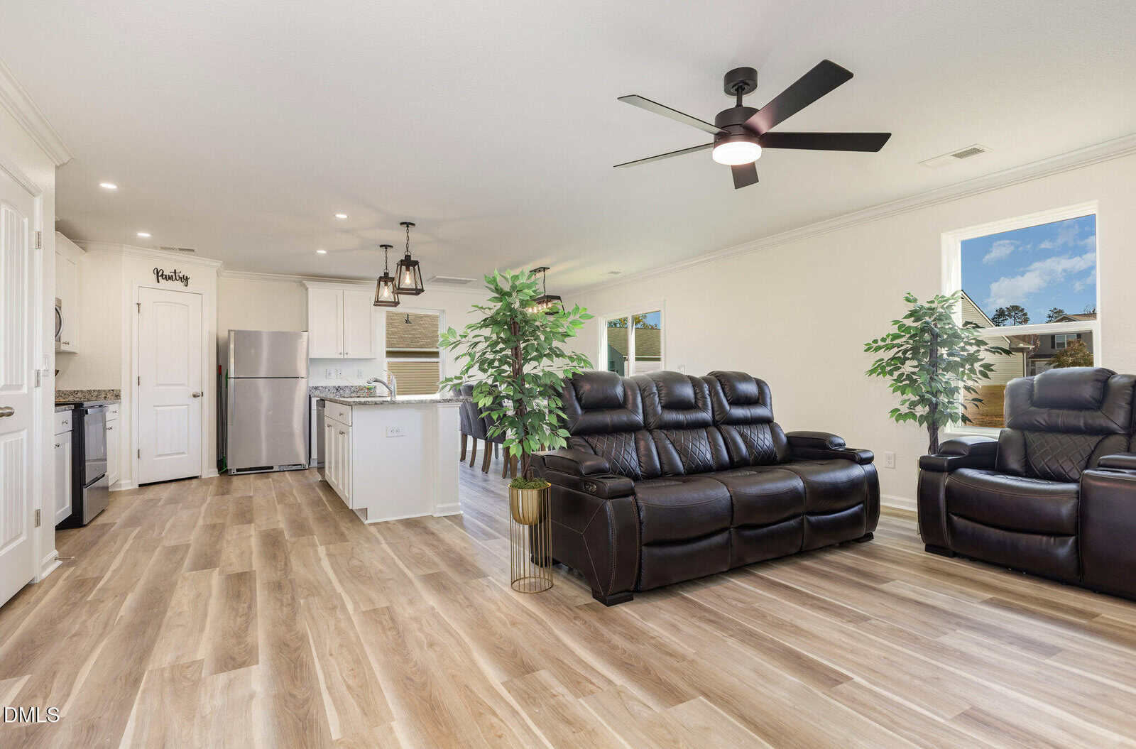 A modern open-concept living area with wood-style floors, black leather reclining sofas, potted plants, and a ceiling fan. The kitchen features white cabinets, stainless steel appliances, and pendant lights over an island.