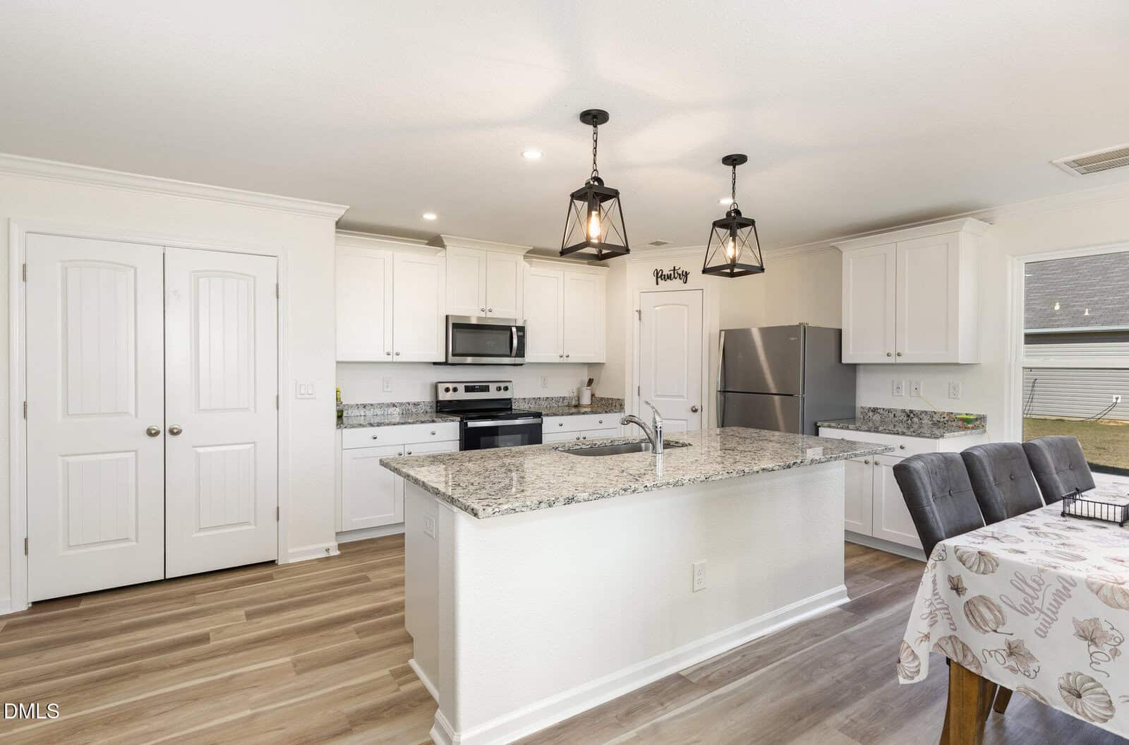 A modern kitchen with white cabinets, stainless steel appliances, a granite island with a sink, pendant lights, and a dining table with a patterned tablecloth; double doors are on the left.