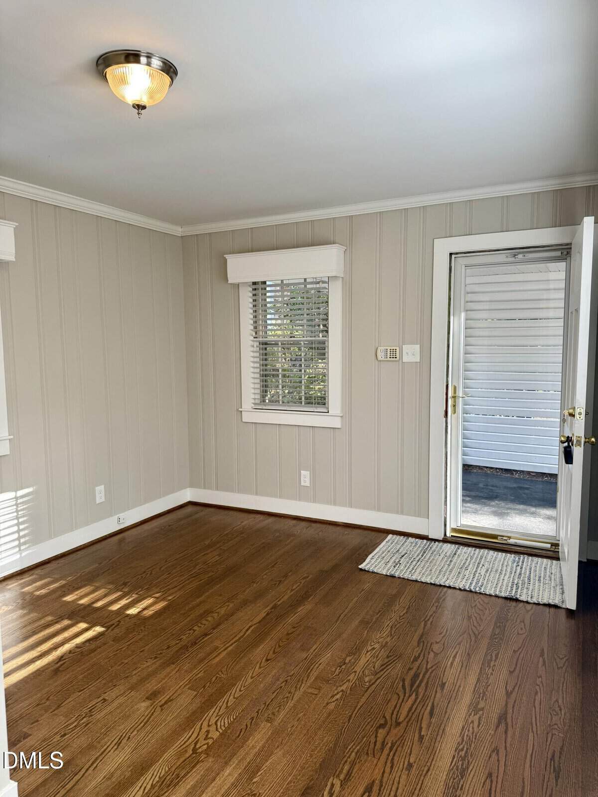 Bright, empty room with wood floors, beige paneled walls, a window with white blinds, and a glass door leading outside. Ceiling light fixture and small rug near the entrance.