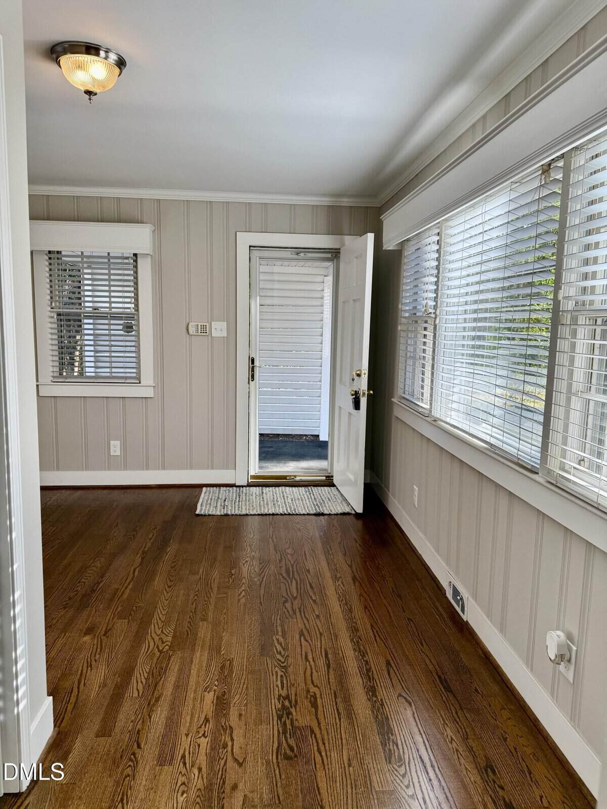 Bright hallway with hardwood floors, light-colored paneled walls, large windows with blinds, and an open door leading to a covered porch area. Ceiling light fixture and a small rug by the entrance.