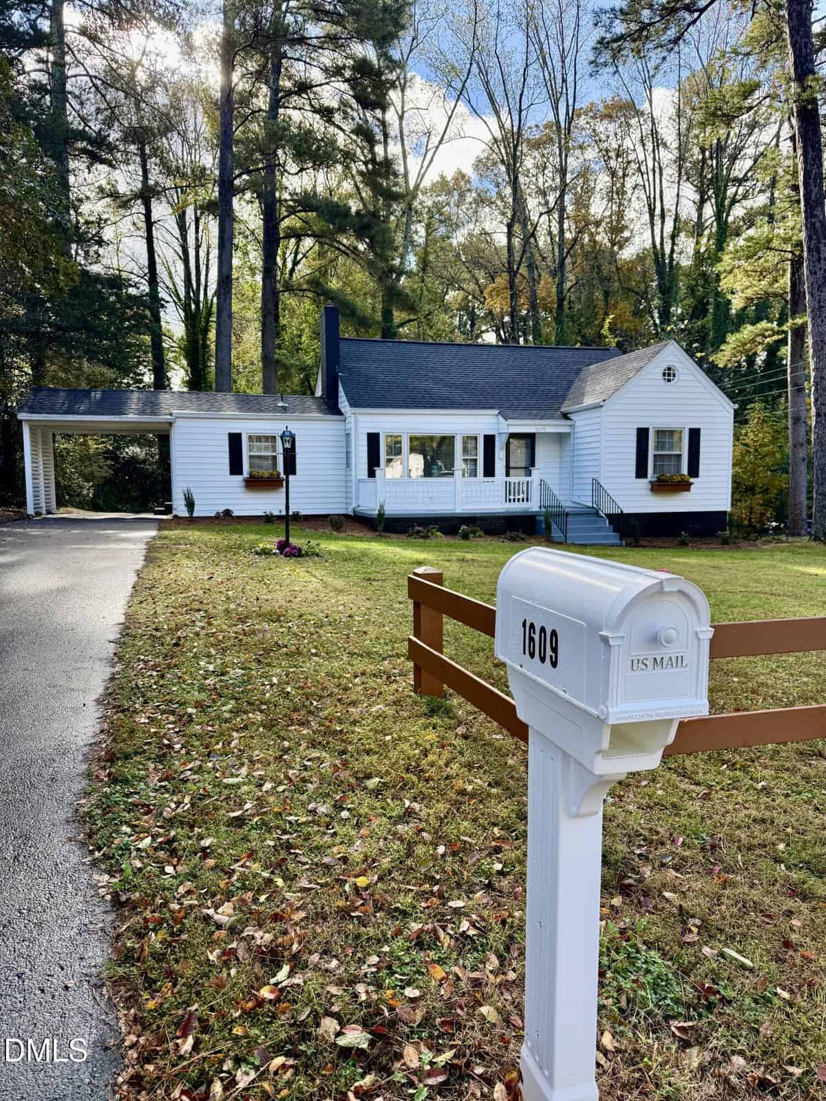 A white mailbox labeled “1609” stands by a wooden fence in front of a white house with a carport, surrounded by tall trees and a neatly mowed lawn.