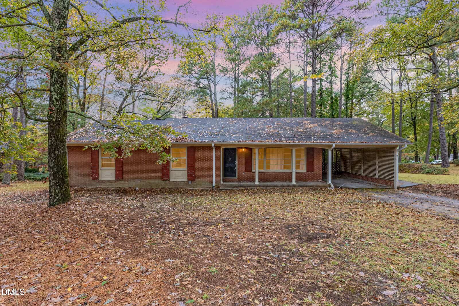 A single-story brick house at 1612 Lynne Avenue in Henderson features a covered porch and attached carport, surrounded by tall trees and fallen leaves beneath a pink and purple sky.