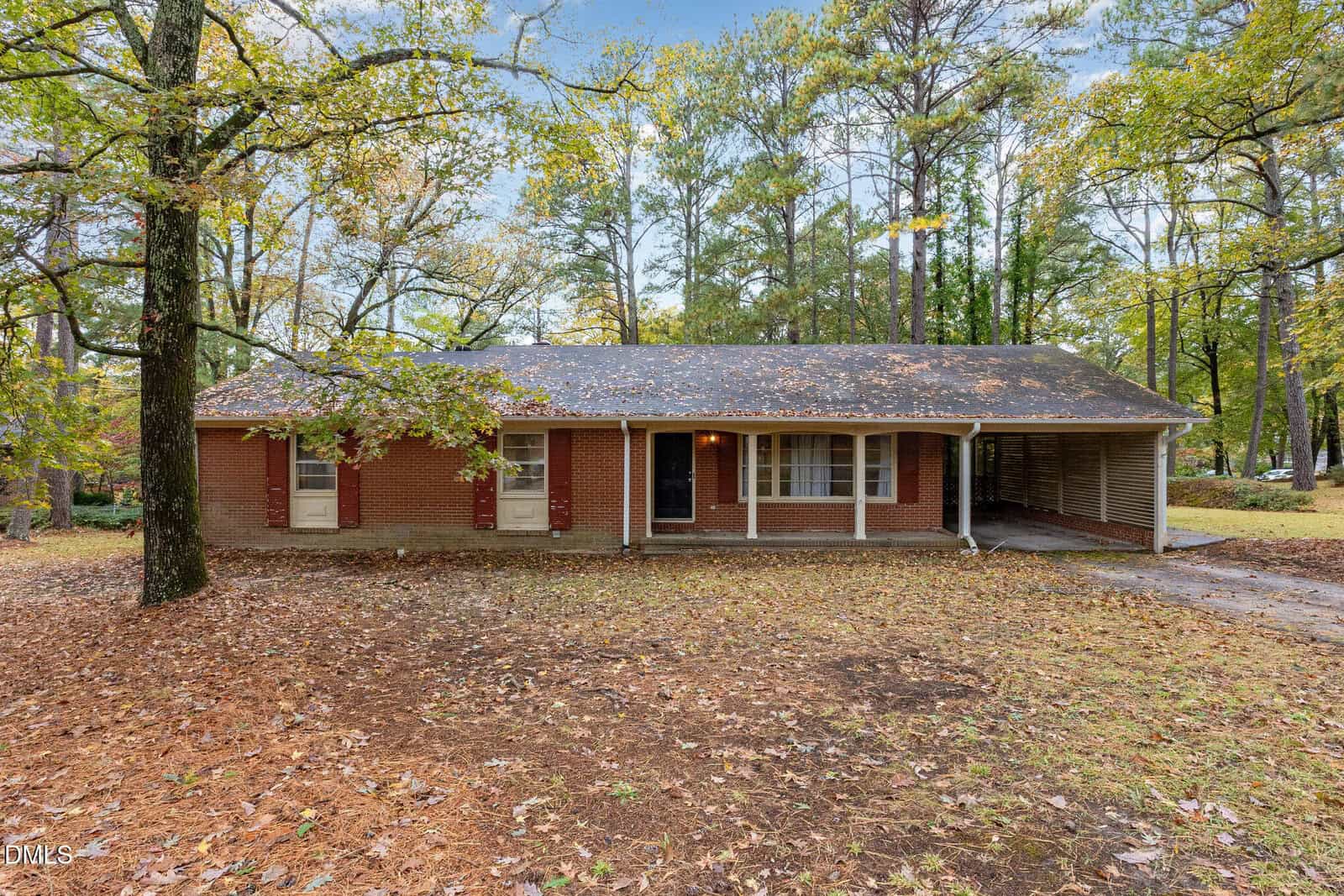 A single-story brick house with a front porch and carport, surrounded by tall trees and fallen leaves on the ground, under an overcast sky.