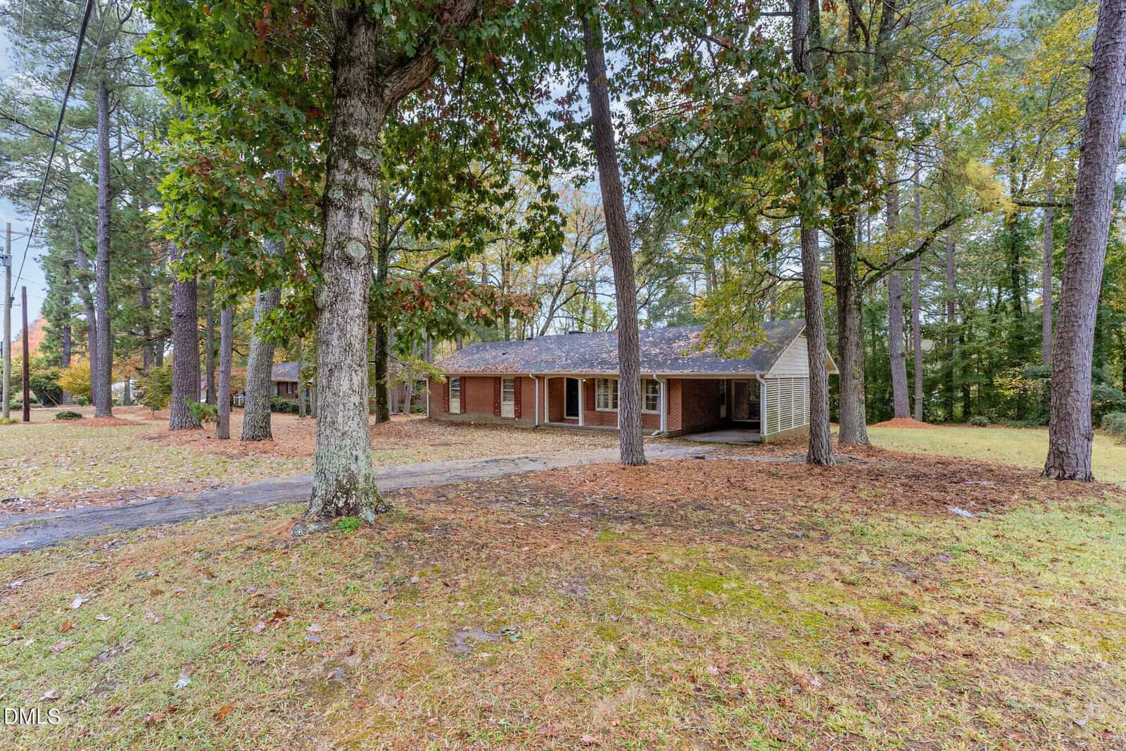 A single-story brick house with a covered front porch, surrounded by tall trees and a large, grassy yard with scattered leaves. A paved driveway leads to the front of the home.