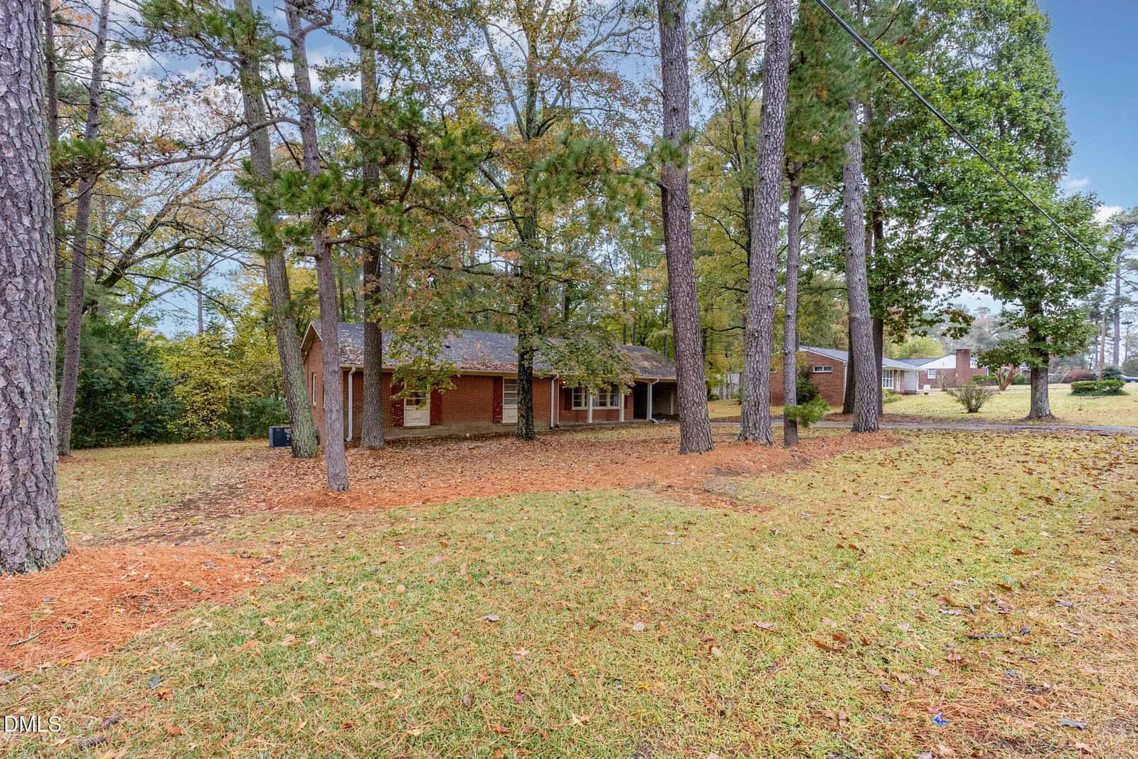 A single-story brick house sits among tall pine trees on a grassy lot. Fallen pine needles and leaves cover the ground, and neighboring houses are visible in the background. The sky is overcast.
