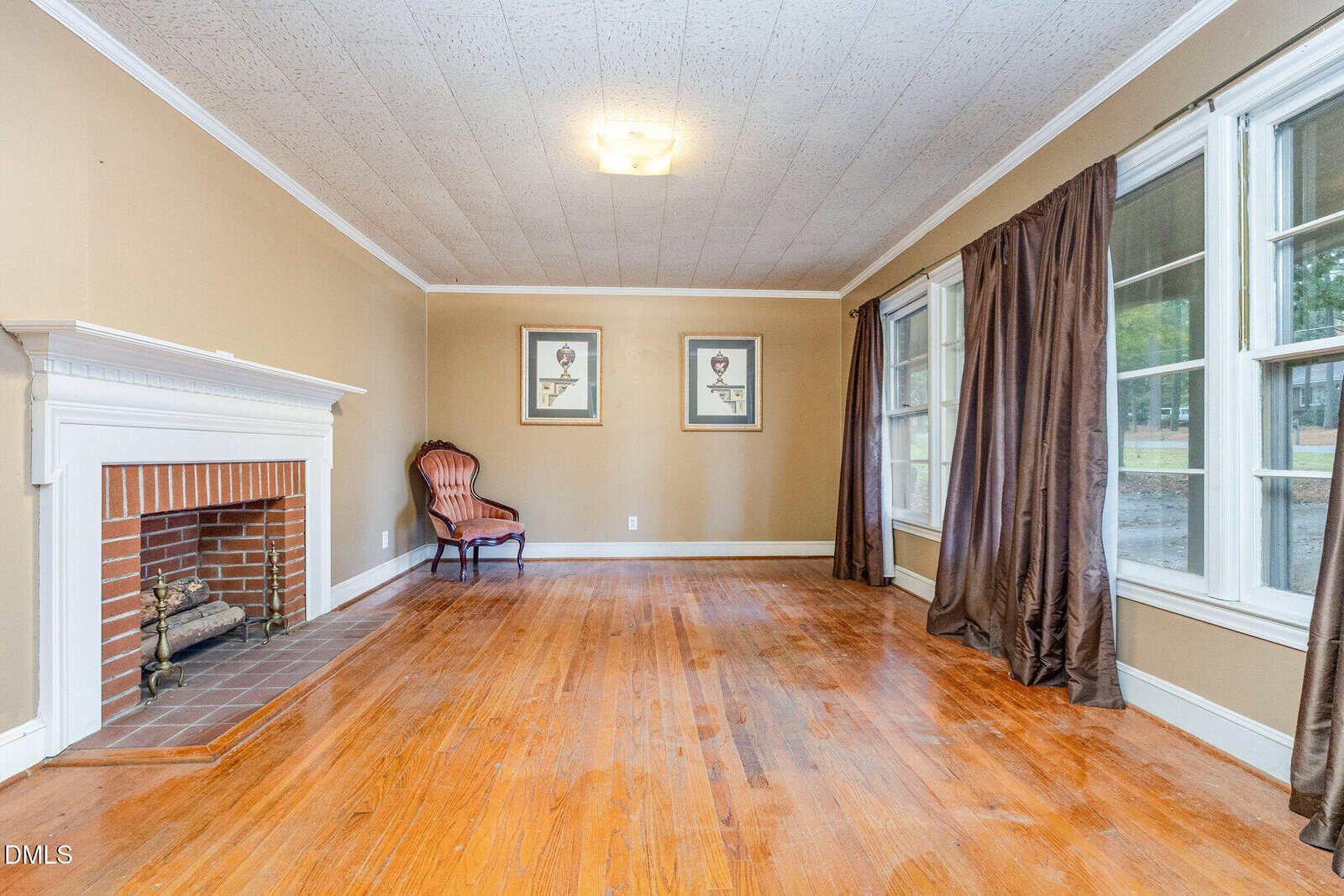 A mostly empty living room with hardwood floors, a white fireplace, a single vintage armchair, two framed pictures on the far wall, and tall windows with brown curtains letting in natural light.