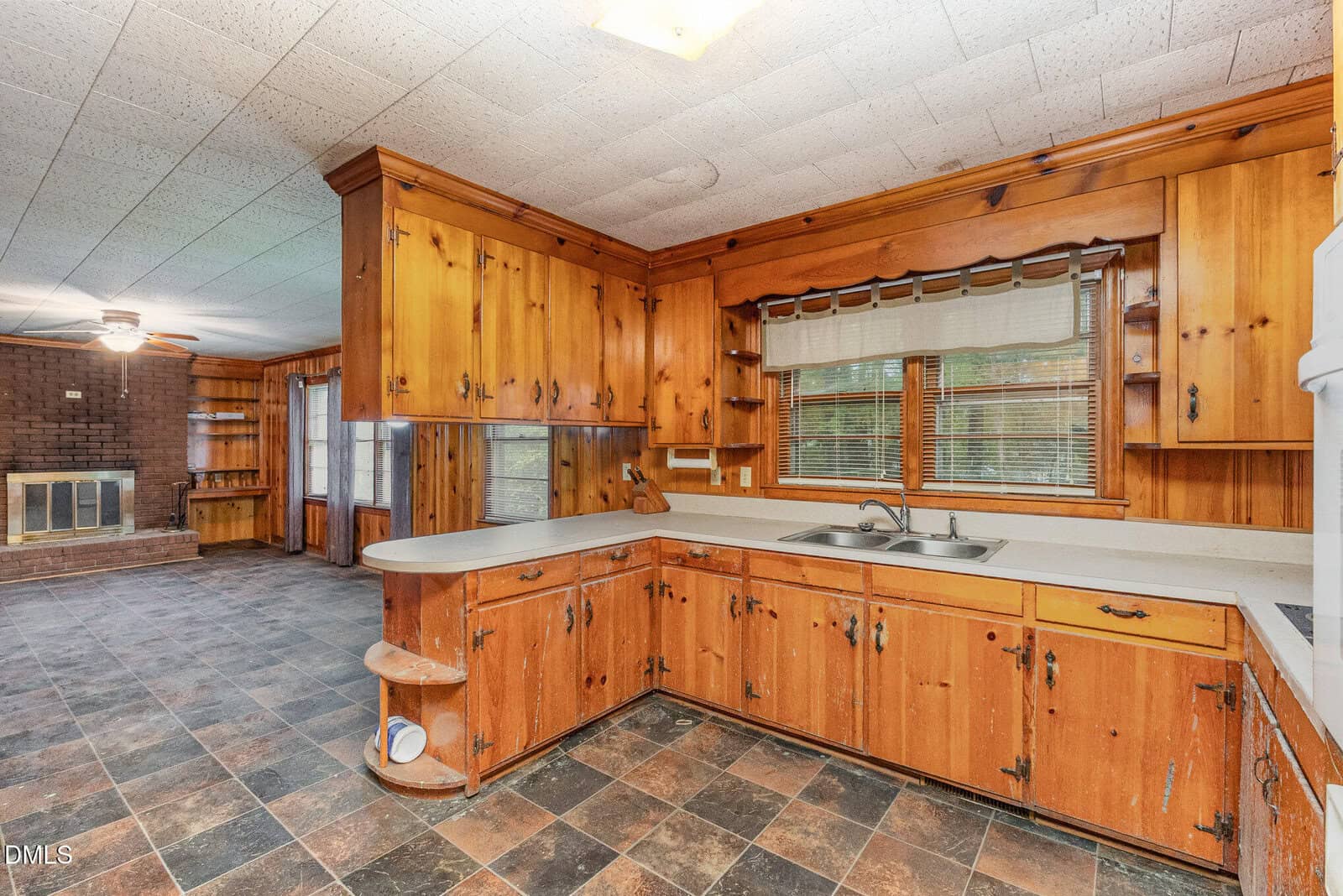 A spacious kitchen with wood-paneled cabinets, a double sink under a window, tiled floors, and open shelving. The adjoining room features a fireplace, wood walls, and large windows.