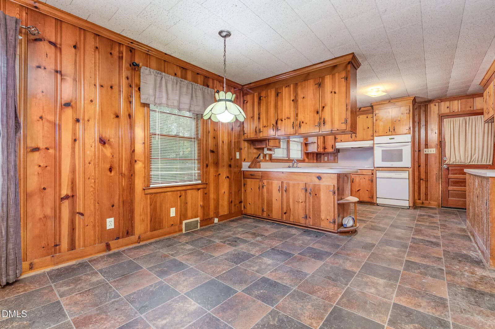 A kitchen and dining area with wood-paneled walls, tile flooring, a hanging light fixture, white appliances, a large window with a curtain, and built-in cabinets and shelving.