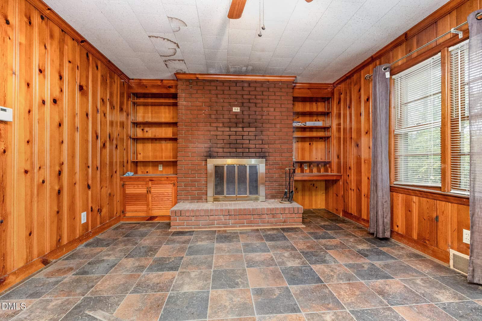 A wood-paneled room with a brick fireplace centered on one wall, flanked by built-in wood shelves and cabinets. The floor is tiled in a dark, mottled pattern. Large windows with curtains are on the right side.