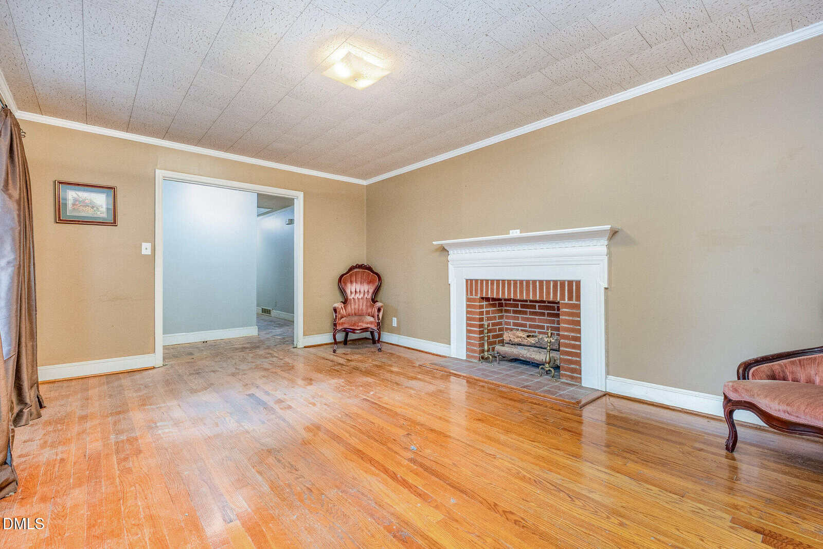 A living room with light brown walls, a wooden floor, a white fireplace, two vintage pink chairs, and a picture on the wall. The room is brightly lit with a ceiling light and has a doorway leading to another area.
