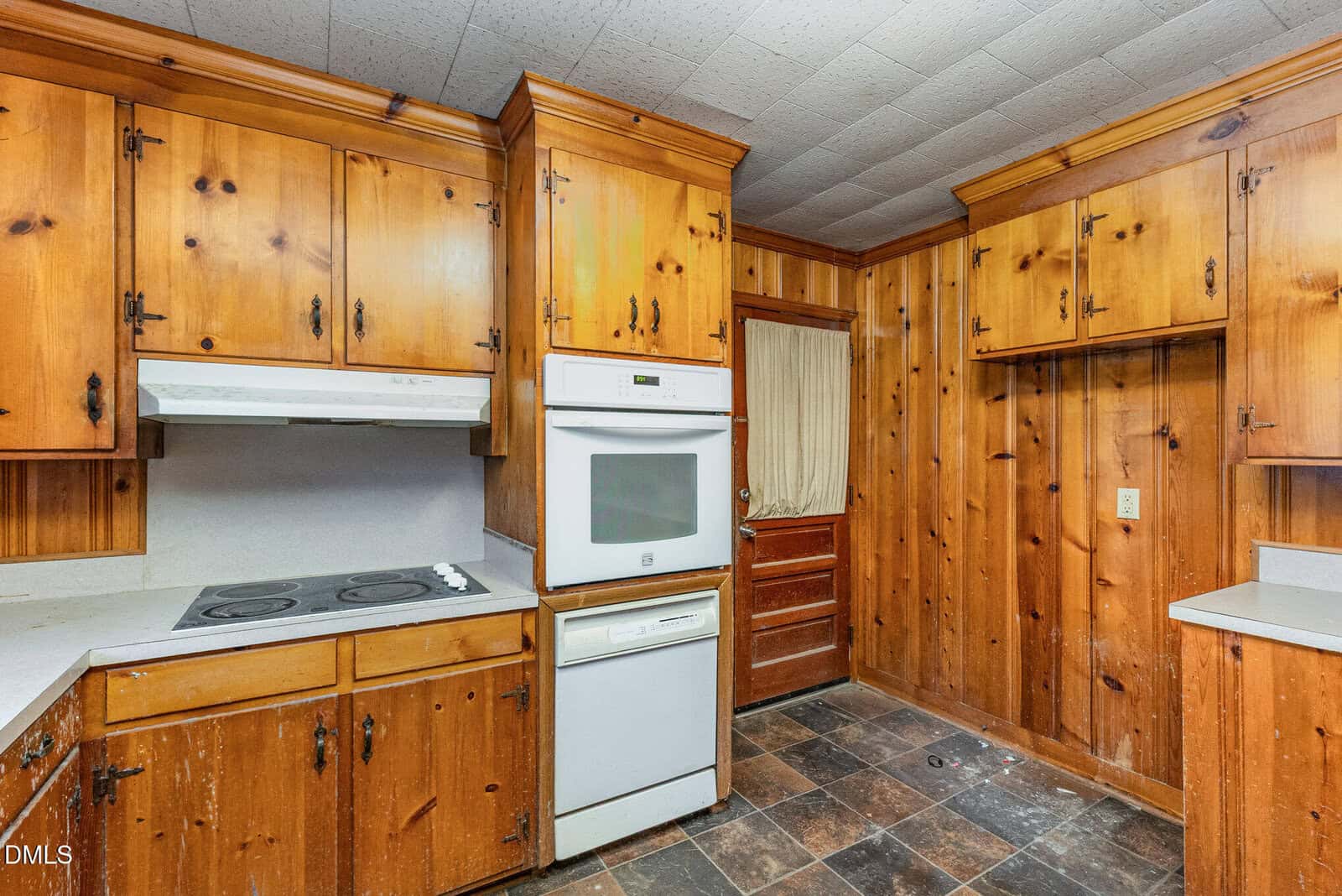A kitchen with wood-paneled cabinets and walls, a white double oven, an electric cooktop with a range hood, and dark tiled flooring. The countertops are light-colored, and there is a wooden door in the background.