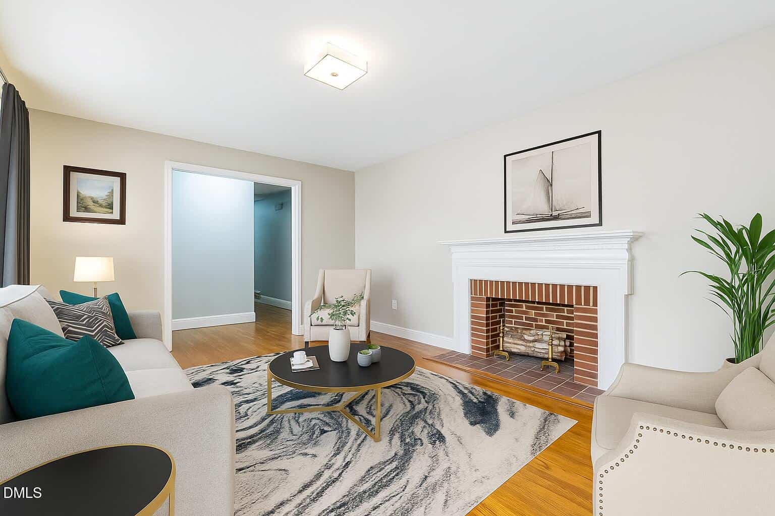 A living room with a brick fireplace, two armchairs, a sofa, a round coffee table, a potted plant, framed artwork, and a patterned rug on a wooden floor. Walls are light-colored and the room is well-lit.