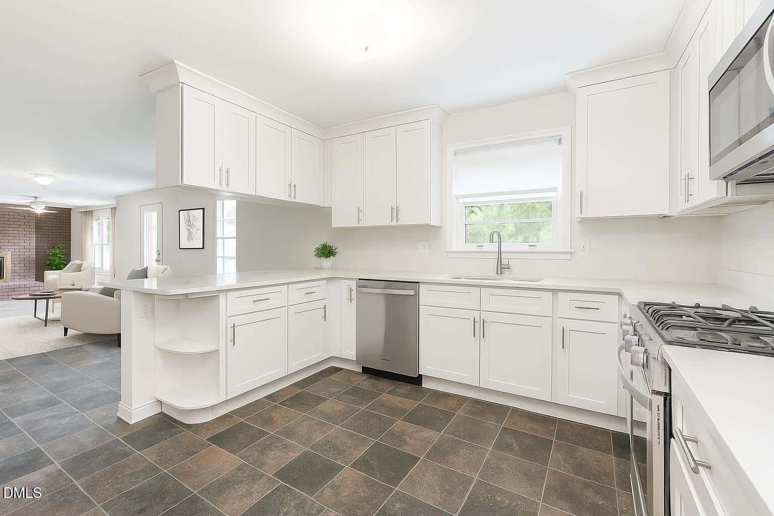 Modern kitchen with white cabinets, stainless steel appliances, a large window above the sink, and dark tile flooring. The kitchen opens to a living area with a brick fireplace visible in the background.