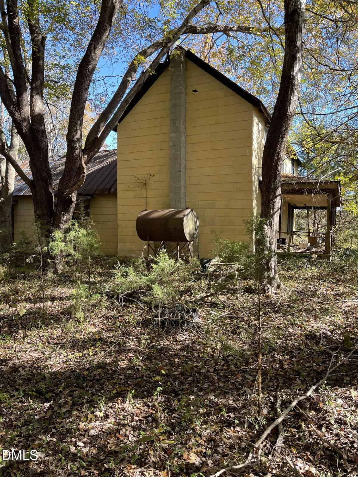 A yellow two-story house with a metal chimney and oil tank in a wooded area. The house appears weathered and overgrown with vegetation, and sunlight filters through the trees.