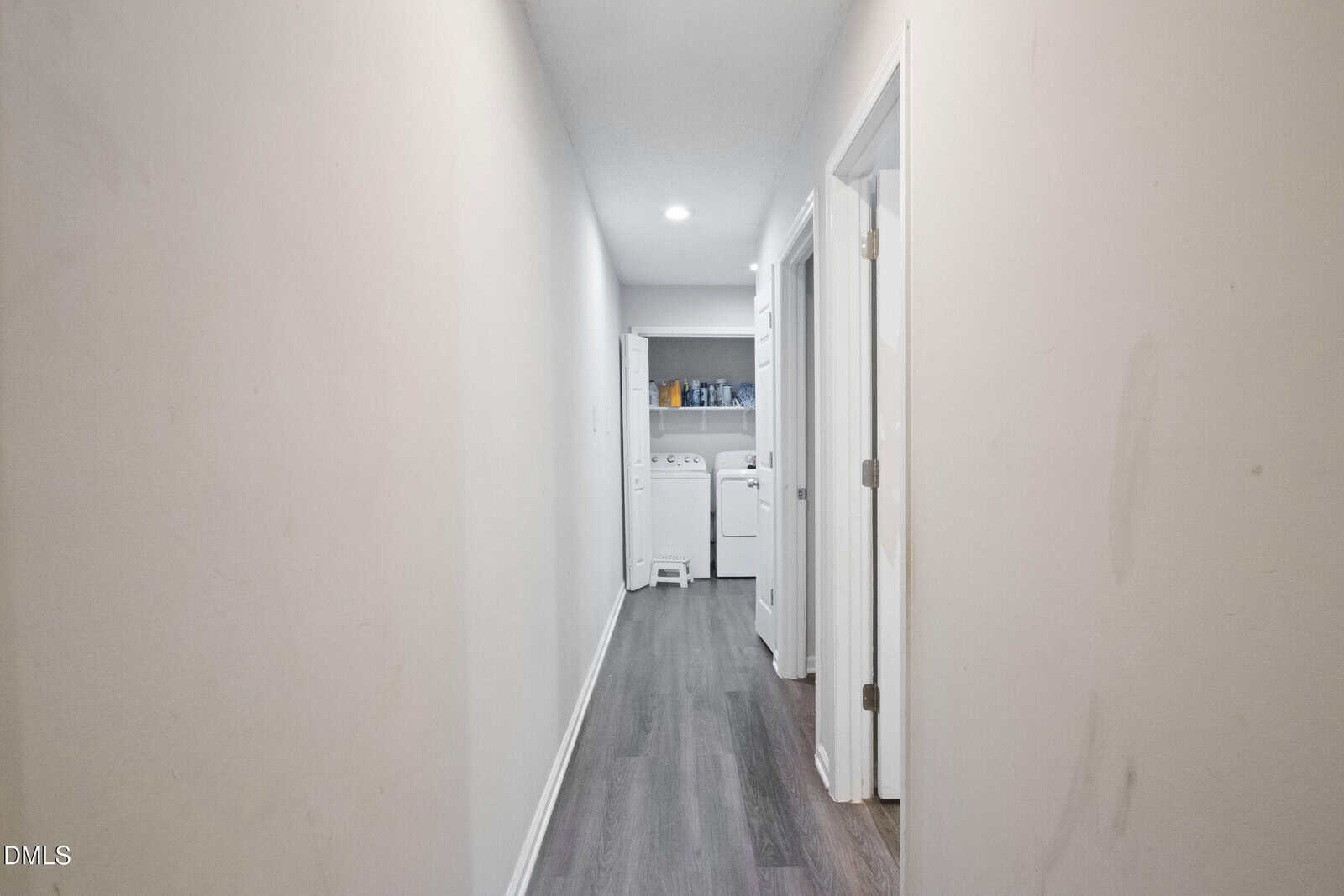 A hallway with light-colored walls and dark flooring leads to a small laundry area with a washer and dryer. There are shelves above the machines holding various items. Two doorways are on the right side of the hallway.