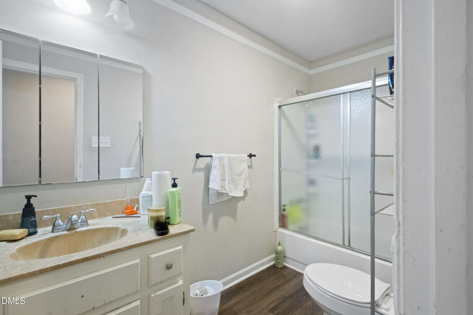 A bathroom with a beige countertop, white sink, and mirror above. Towels hang on a black towel rack. There is a white toilet, shelving unit, frosted glass shower door, and wood-style flooring. Toiletries are on the counter.