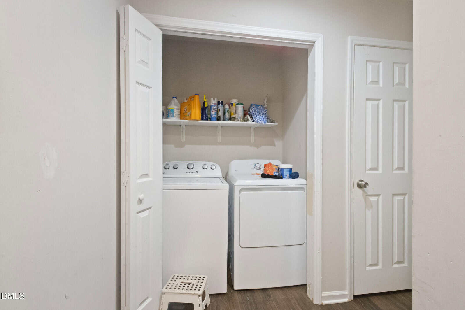 A laundry area with a washing machine and dryer beneath a white shelf holding detergent and cleaning supplies. The space is enclosed by open double doors, with a small white step stool in front.
