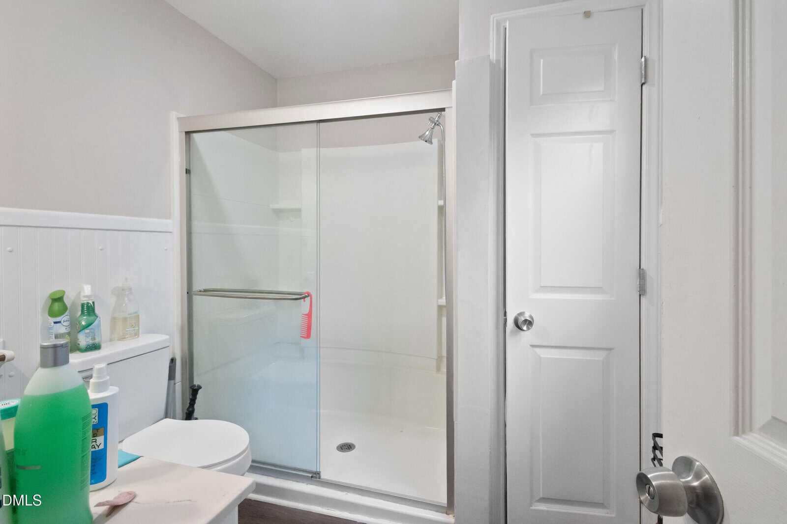 A bathroom with a glass-enclosed shower, a white toilet, cleaning supplies on the toilet tank, and a closed white door. The walls are light-colored and the floor is dark.