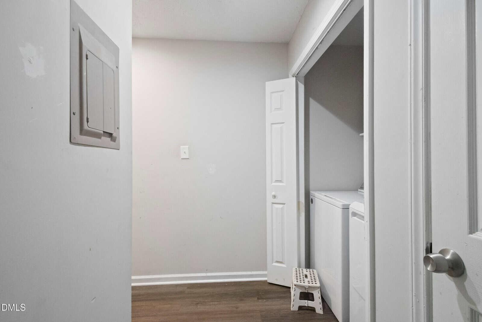 A small laundry area with a top-loading washing machine, a small white step stool, wood-look flooring, a wall-mounted electrical panel, and a partially open closet door. The walls are plain and light-colored.