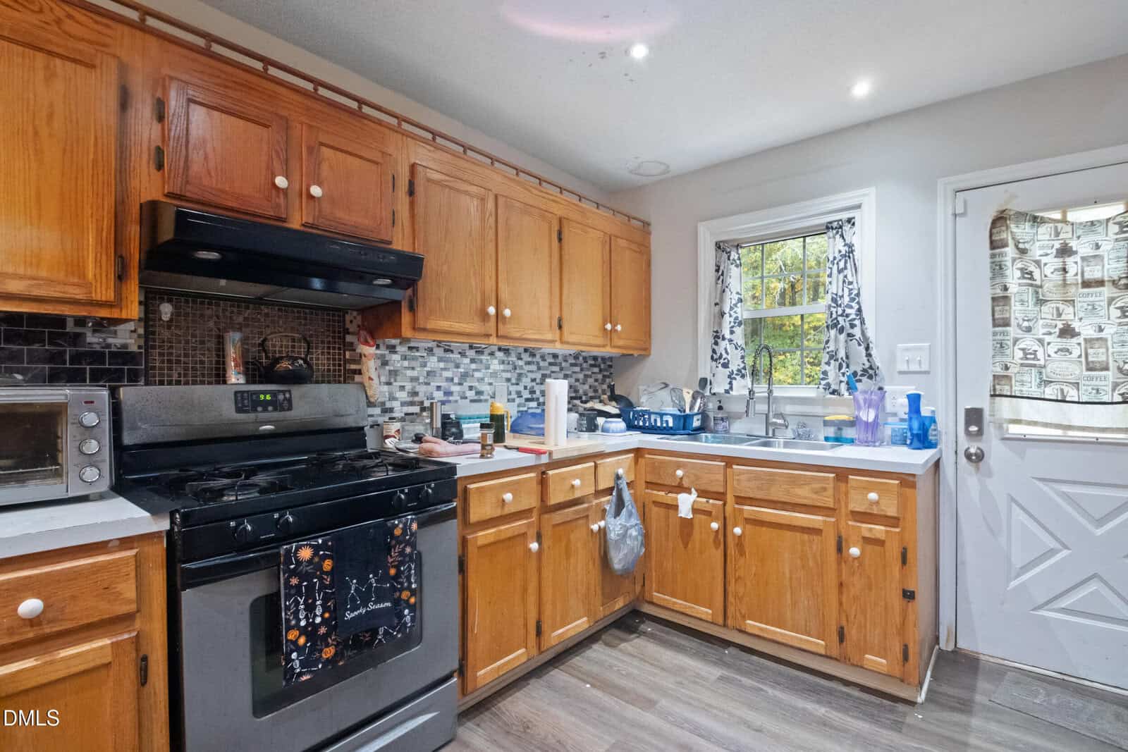 A kitchen with wooden cabinets, a black stove and oven, a microwave, various utensils and containers on the countertops, a window above the sink, and a door with a patterned curtain. The floor is dark wood.
