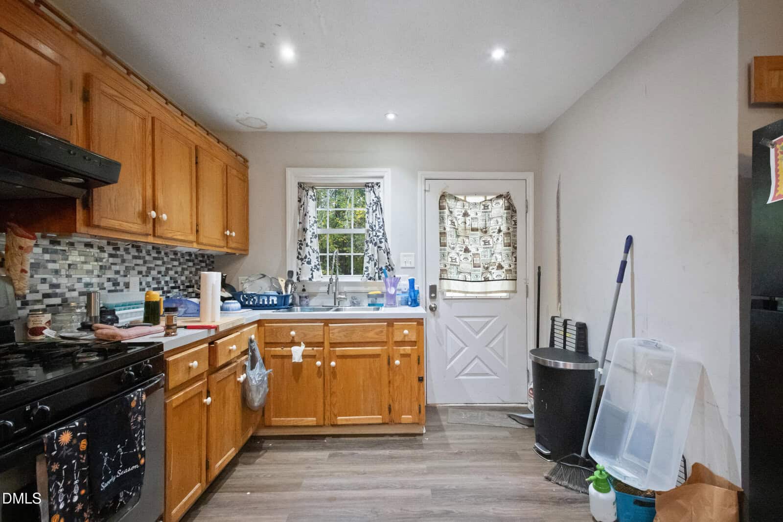 A small kitchen with wooden cabinets, a black stove, a sink under a window with patterned curtains, a white door, a mop, a trash can, and various cleaning supplies and items on the counter and floor.