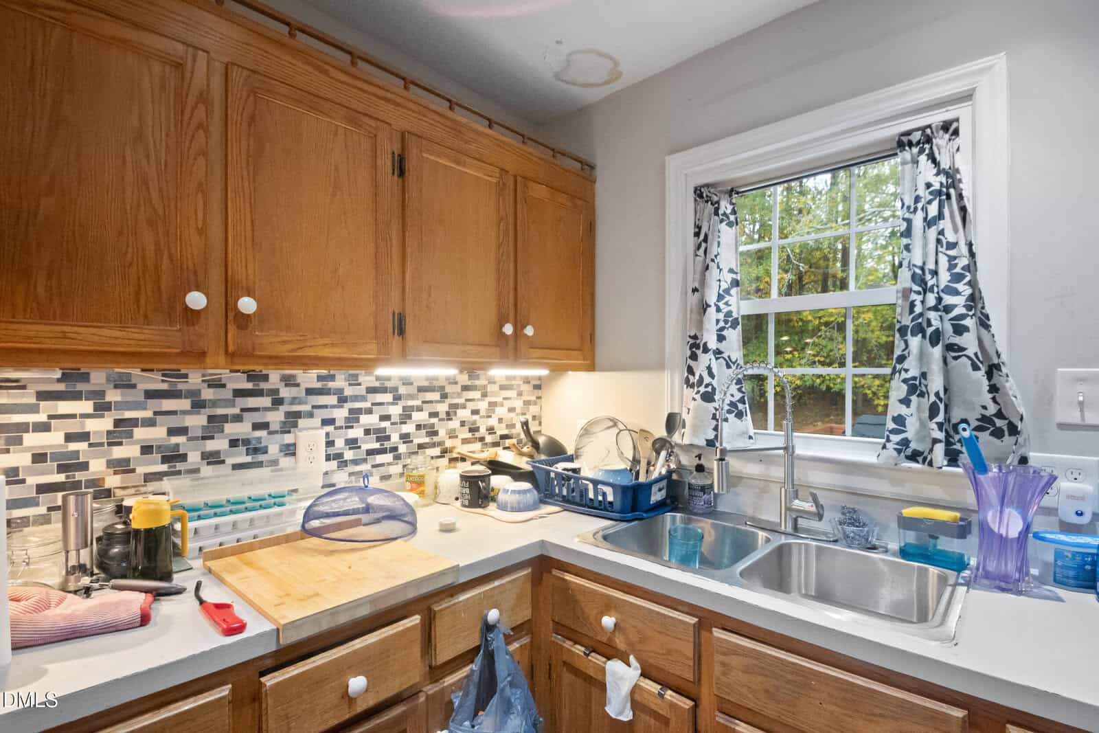A kitchen with wooden cabinets, a tiled backsplash, and a double sink under a window with floral curtains. Countertops hold various dishes, a cutting board, and utensils. A water stain is visible on the ceiling.