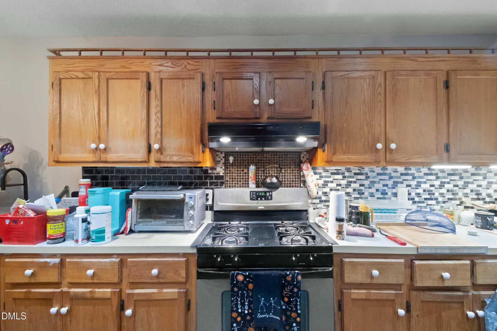 A kitchen with wooden cabinets, a black and stainless steel gas stove, a black range hood, a mosaic tile backsplash, a toaster oven, various kitchen items, and utensils on the counter.