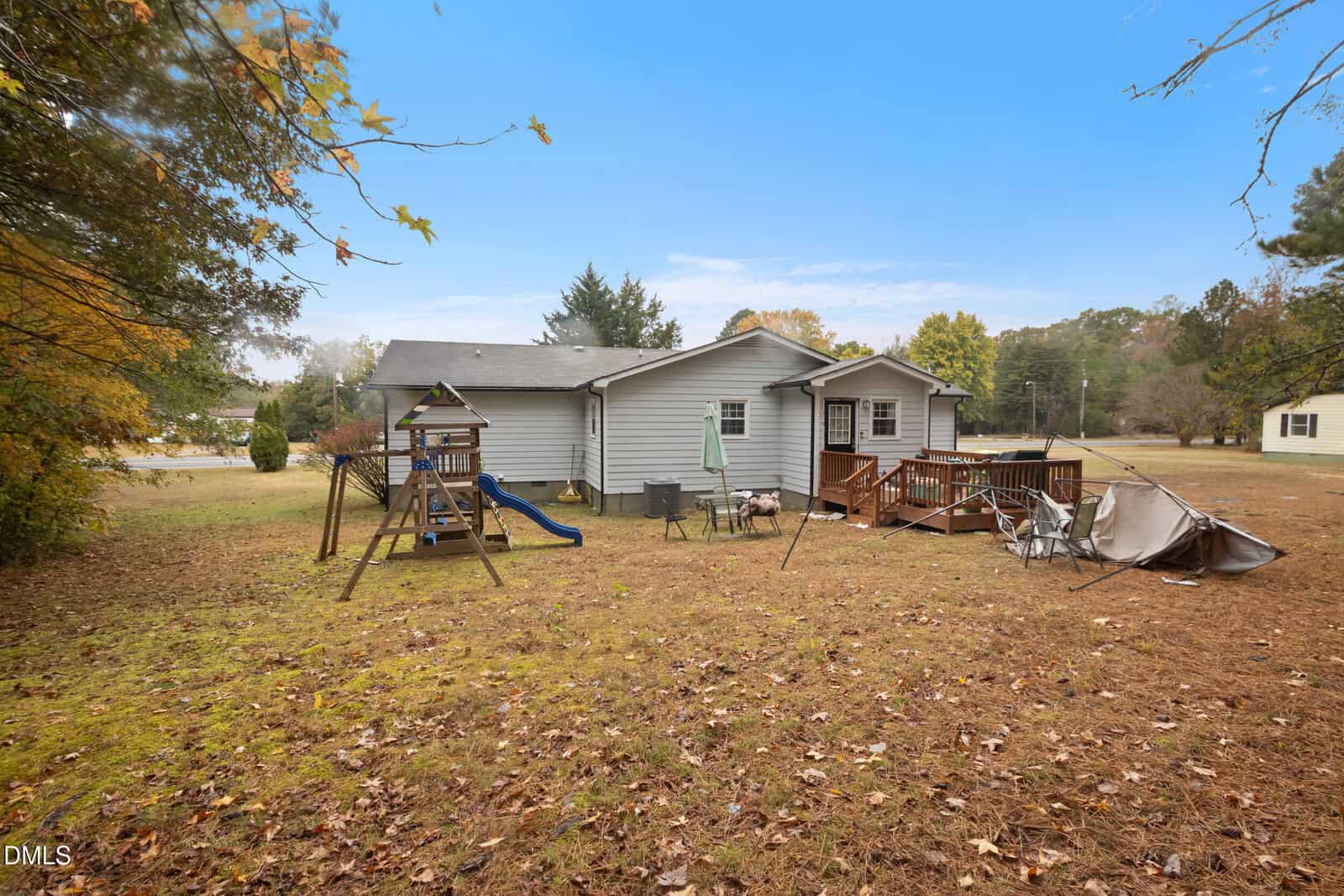A gray house with a backyard, wooden playset with a slide, patio umbrella, small deck with chairs, and a collapsed canopy on a grassy lawn surrounded by trees.