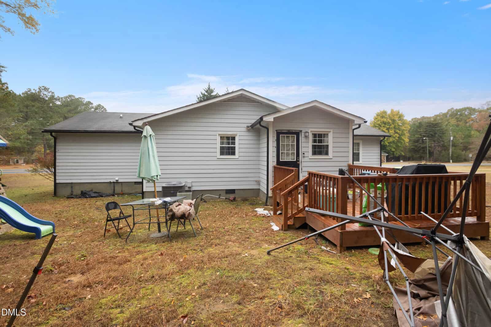 A small white house with a wooden deck, outdoor table and chairs, umbrella, and grill in a grassy yard. Trees and blue sky are in the background.