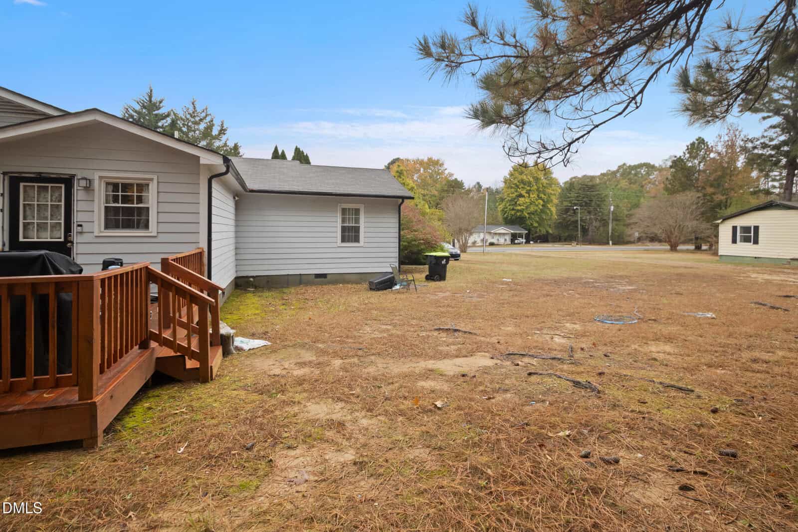 A backyard with dry grass, a wooden deck attached to a gray house, trash bins near the house, and distant homes and trees under a blue sky.