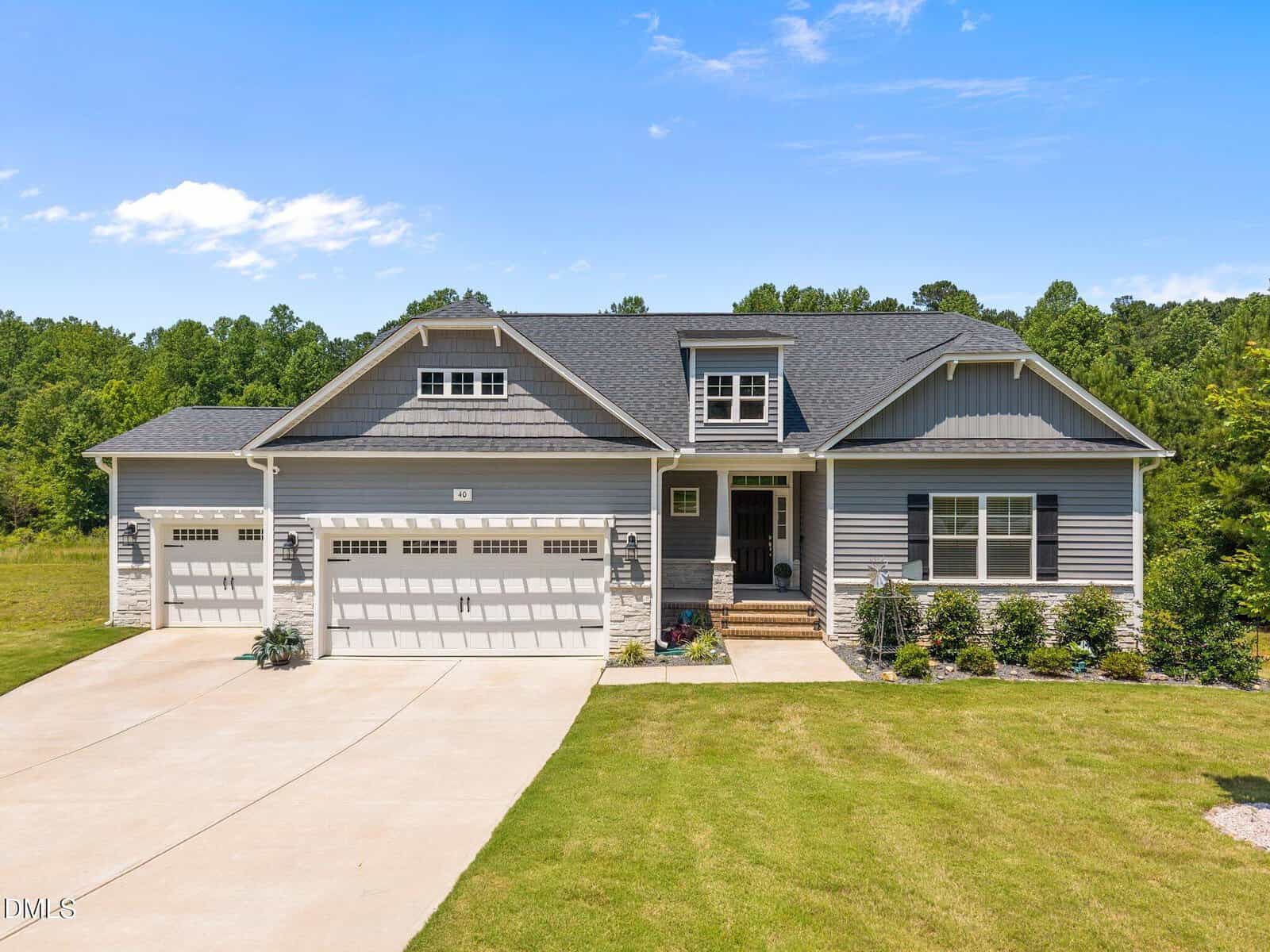 A modern, single-story gray house with stone accents, a three-car garage, front porch, and neatly landscaped yard, set against a backdrop of green trees under a blue sky with scattered clouds.