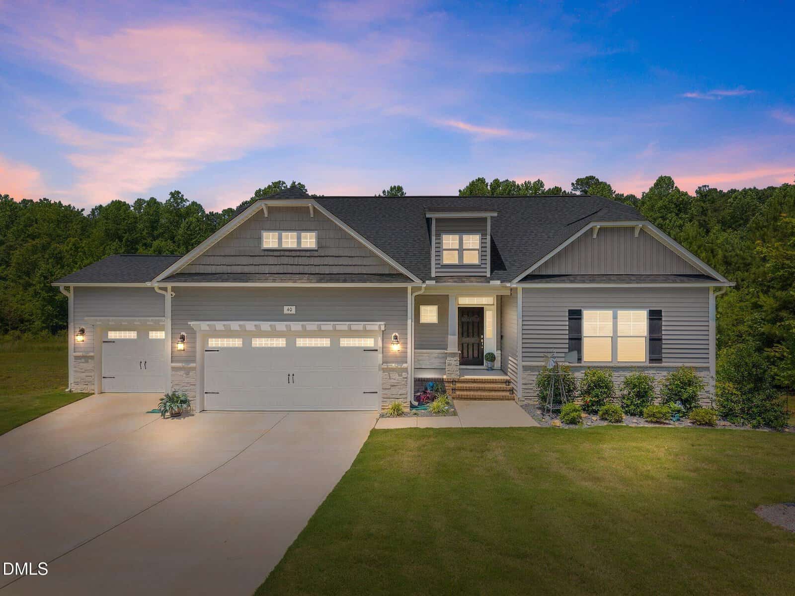 A modern single-story house with gray siding, three garage doors, a manicured front lawn, and porch lights on at dusk, set against a backdrop of trees and a pink and blue sunset sky.