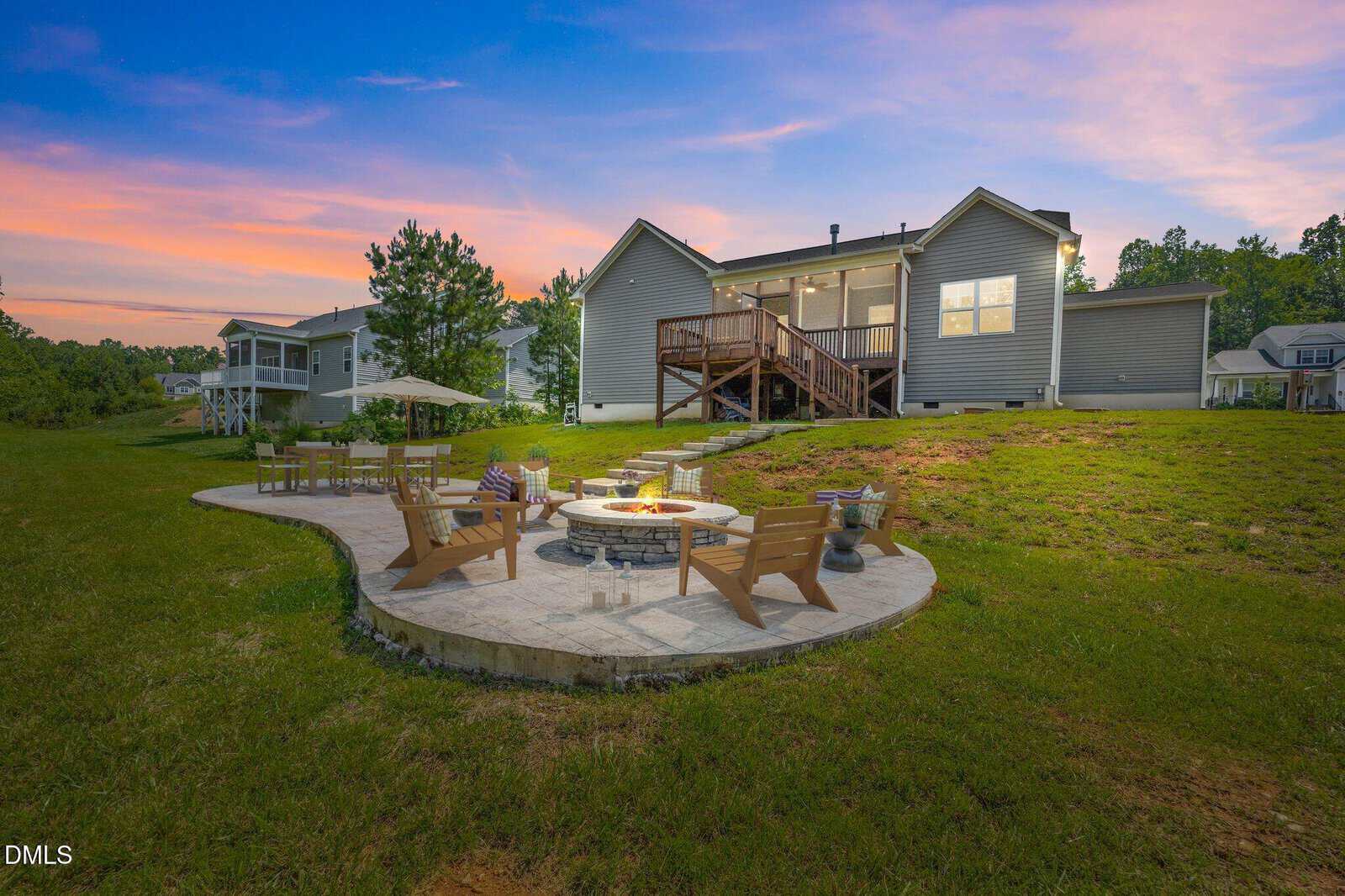 A backyard with a circular stone patio featuring a fire pit, wooden chairs, and small planters. Behind it is a gray house with a raised deck, set against a sunset sky and surrounded by grass and trees.