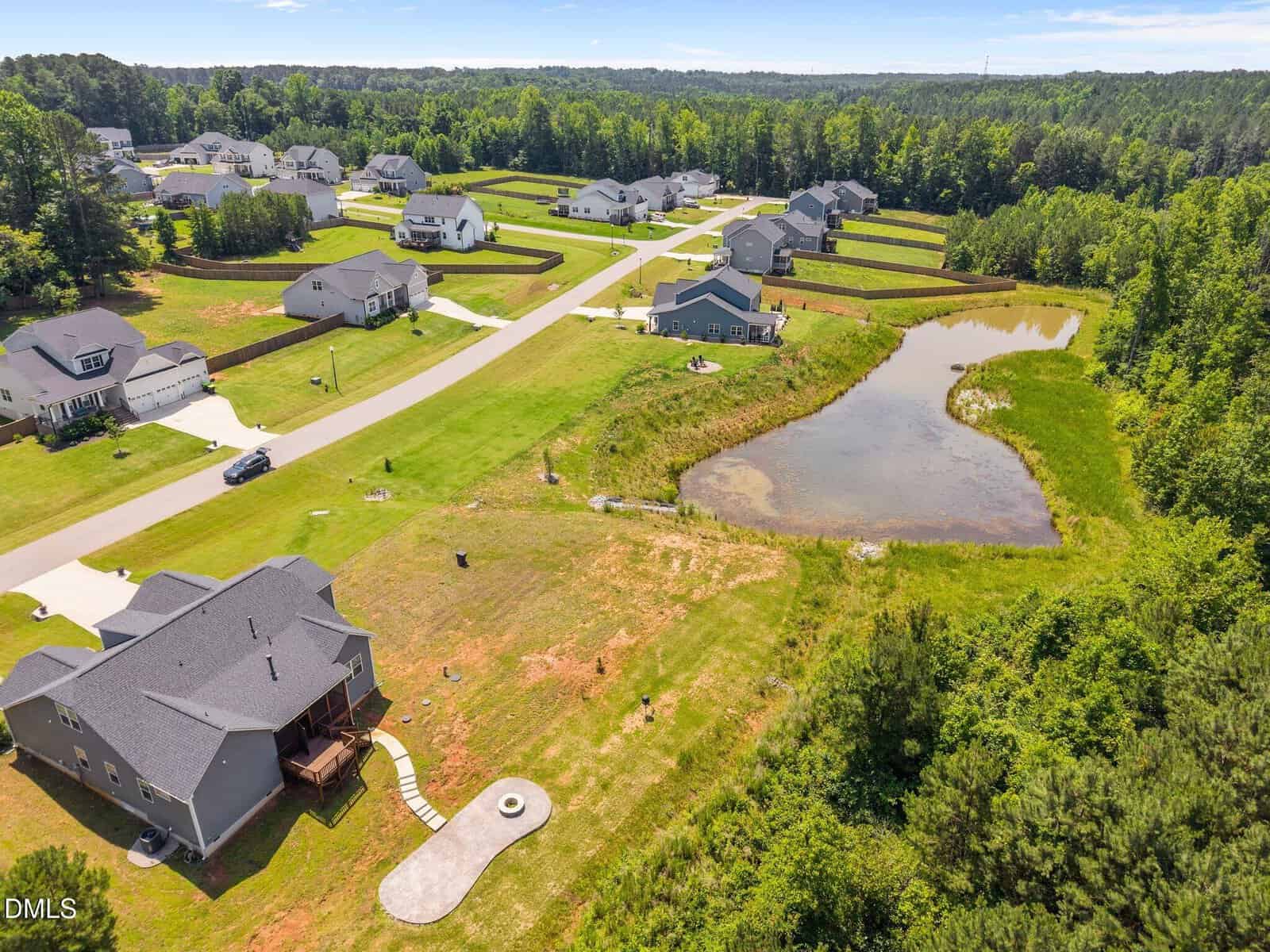 Aerial view of a suburban neighborhood with single-family homes, green lawns, a small pond, and surrounding trees on a sunny day. A winding road runs through the community.