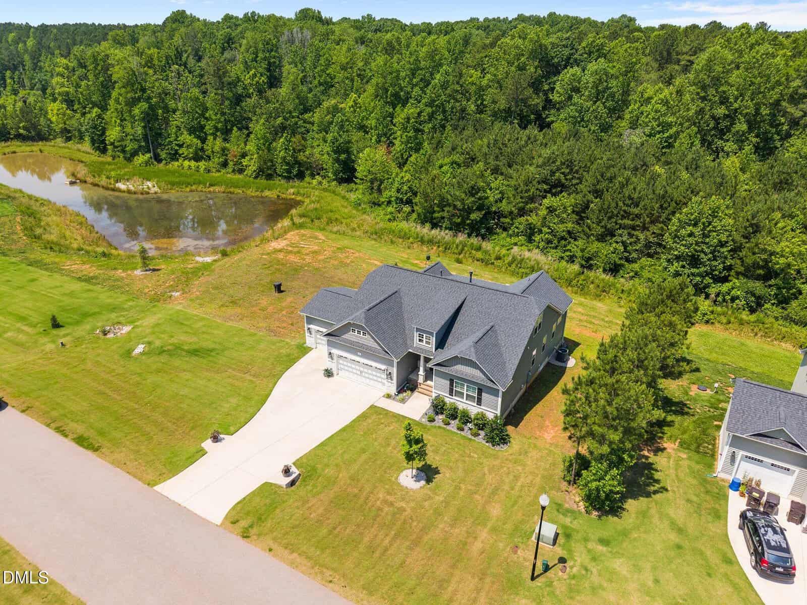 Aerial view of a modern two-story house with a gray roof, attached garage, and large driveway, surrounded by a green lawn, trees, a small pond, and a neighboring house nearby.