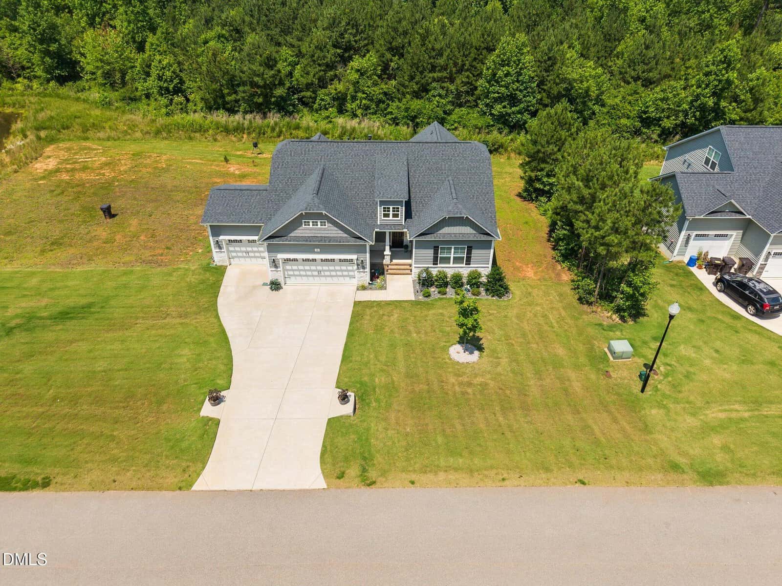 Aerial view of a modern gray house with a dark roof, attached garage, and a neatly manicured lawn. The driveway curves slightly, and the property is bordered by trees and greenery in the background.