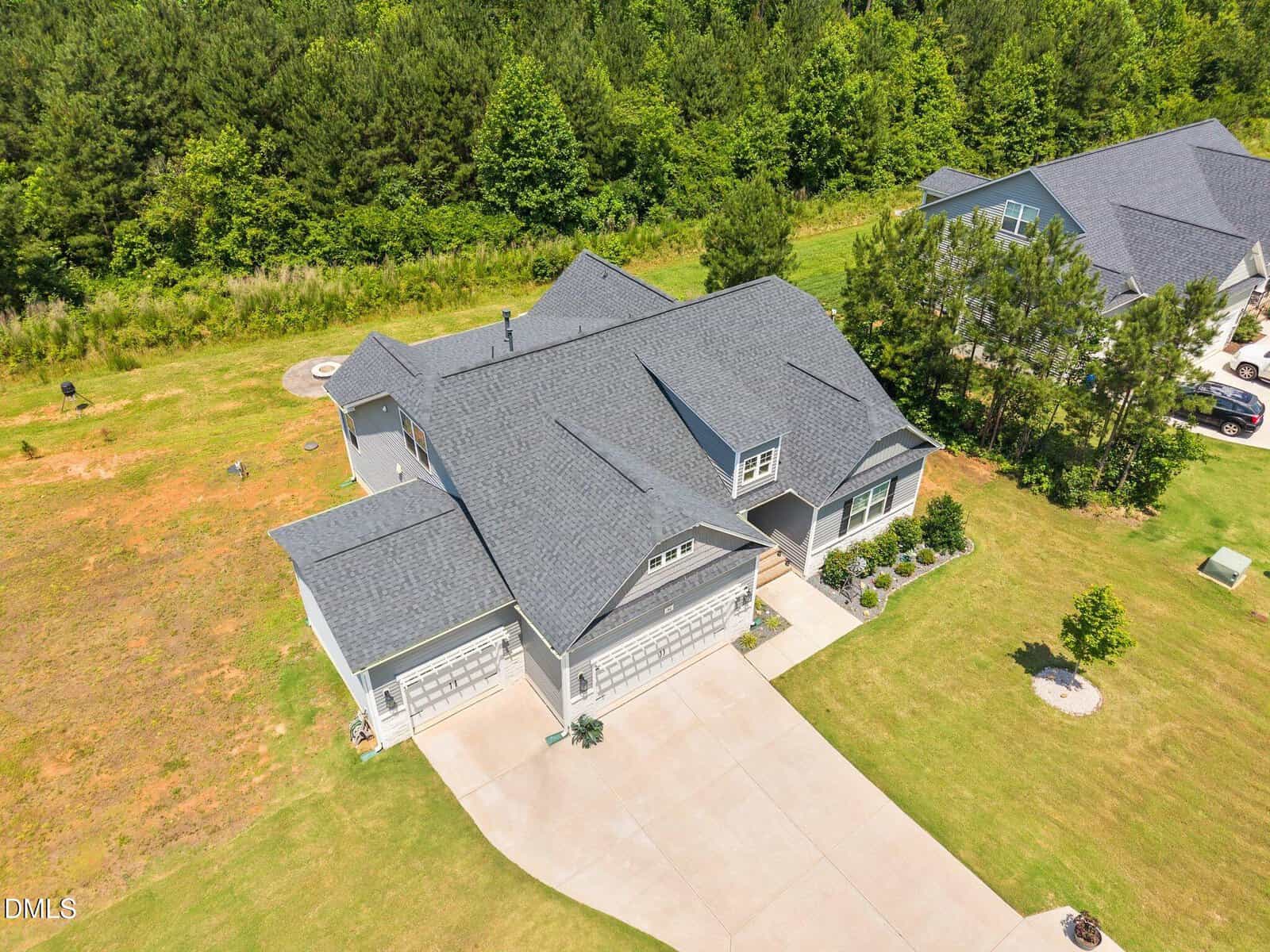 Aerial view of a large, modern gray house with a three-car garage, surrounded by green lawn and trees, with a driveway and a neighboring house visible.