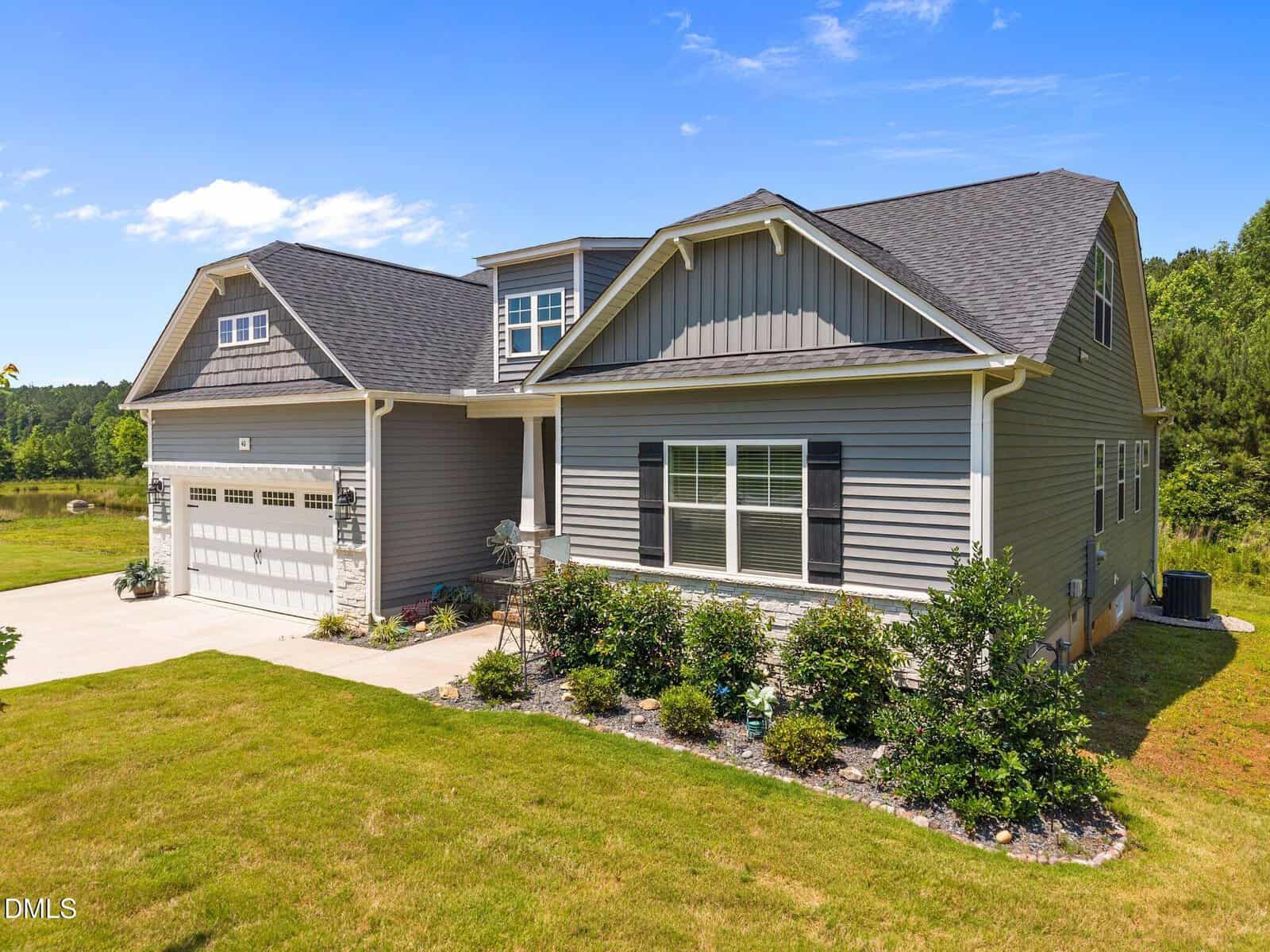 A modern gray two-story house with white trim, a double garage, large windows, and landscaped front yard, set against a backdrop of green trees and blue sky.