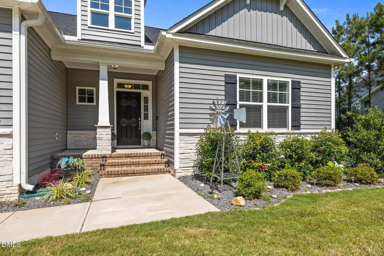 A modern house with gray siding, white trim, a covered entryway with brick steps, landscaped shrubs, and a metal windmill garden decoration next to the front walkway.