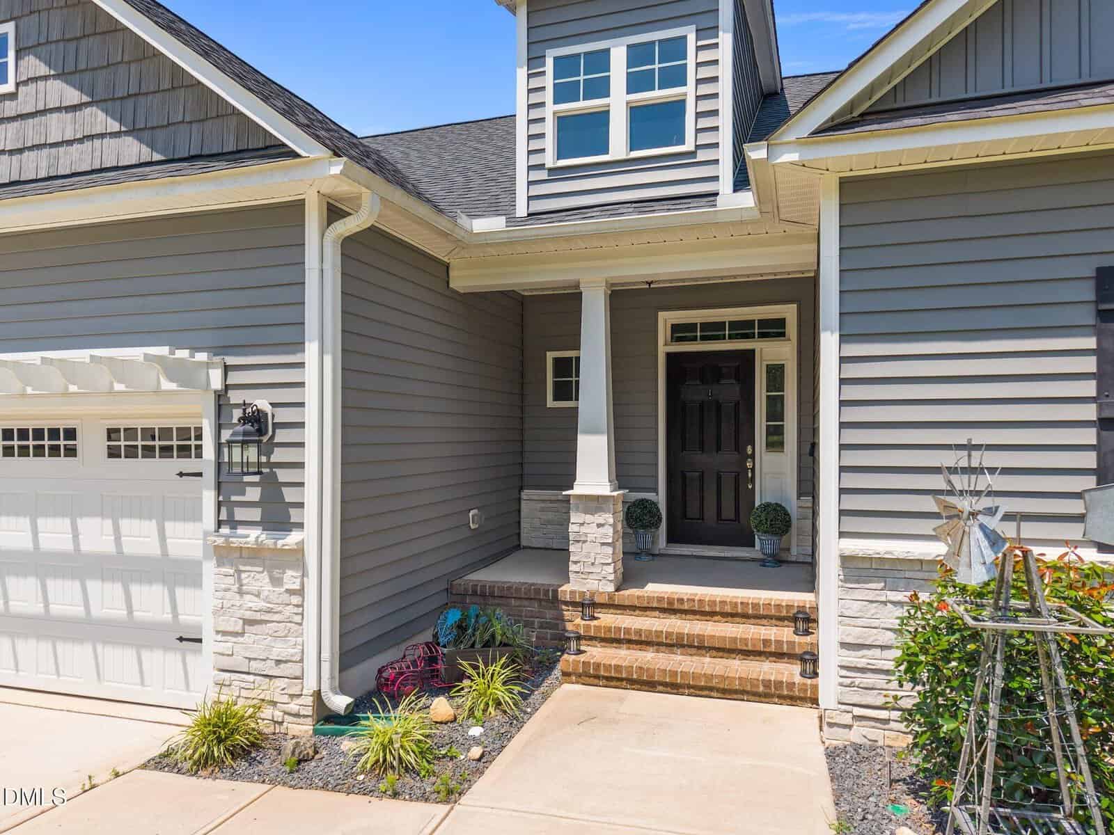 Gray suburban house exterior with white trim, front porch with columns, brick steps, landscaped garden, garage door on the left, and decorative plants along the walkway under a clear blue sky.