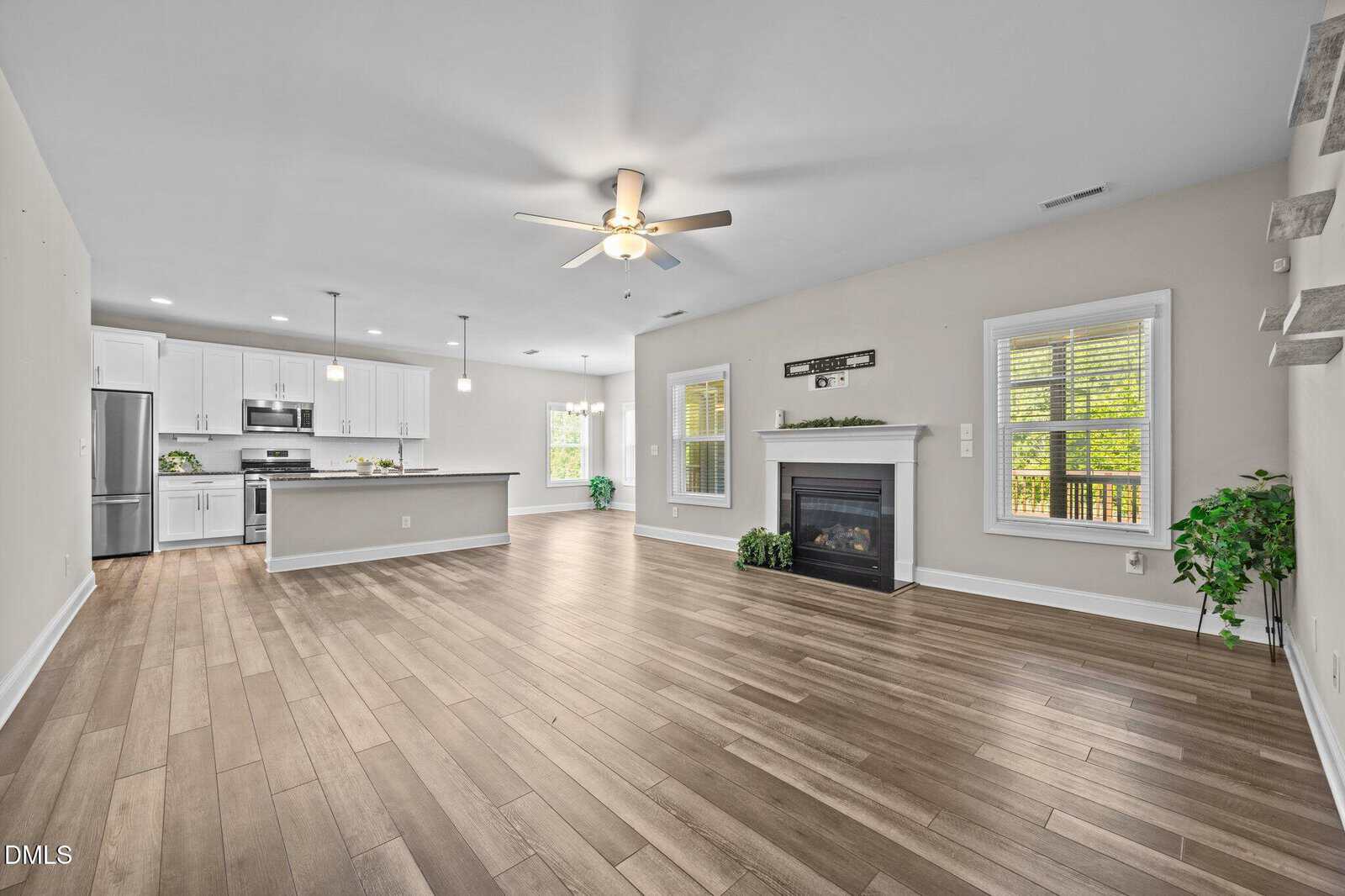 Open-concept living room and kitchen with wood floors, white cabinets, stainless steel appliances, ceiling fan, fireplace, and large windows letting in natural light.