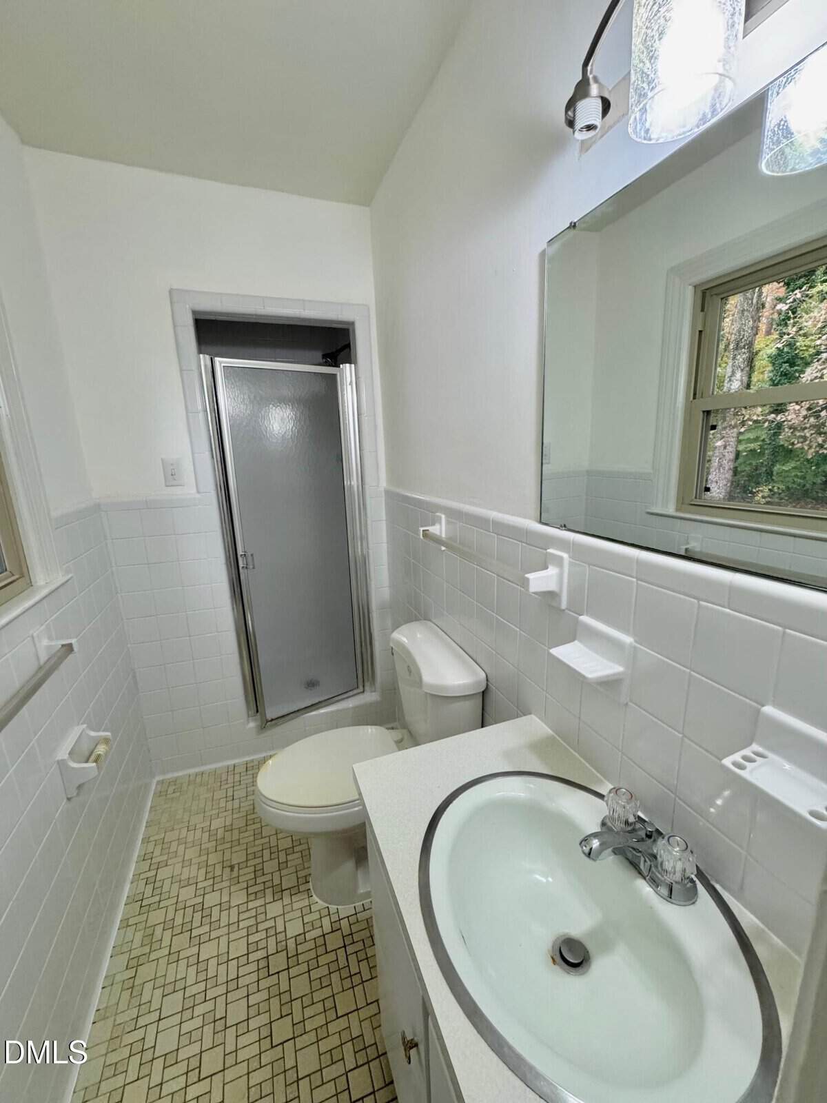 Small bathroom with white tile walls, a toilet, a round sink, a wall mirror, and a glass shower stall. There is a window above the toilet showing green trees outside. The floor has a beige tile pattern.