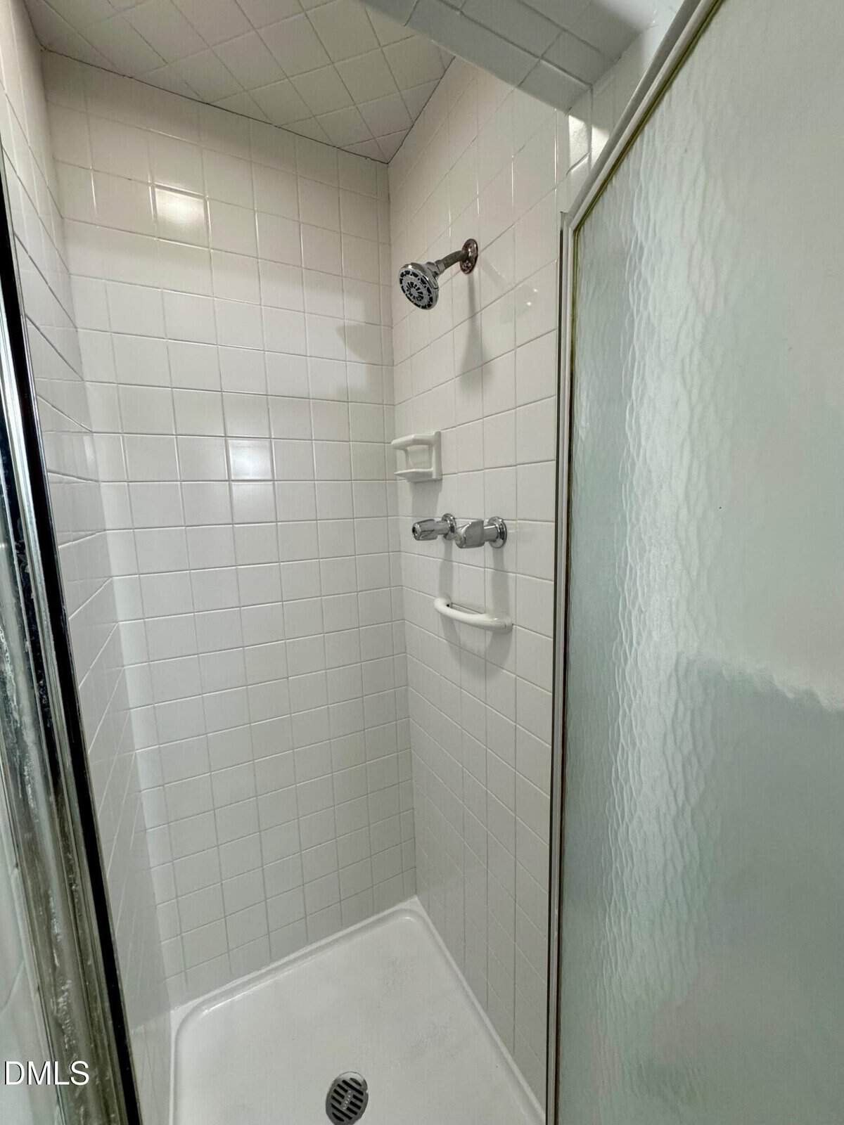 Small, white-tiled shower with a metal showerhead, two built-in soap holders, and a glass door. The floor is white with a round metal drain in the center.