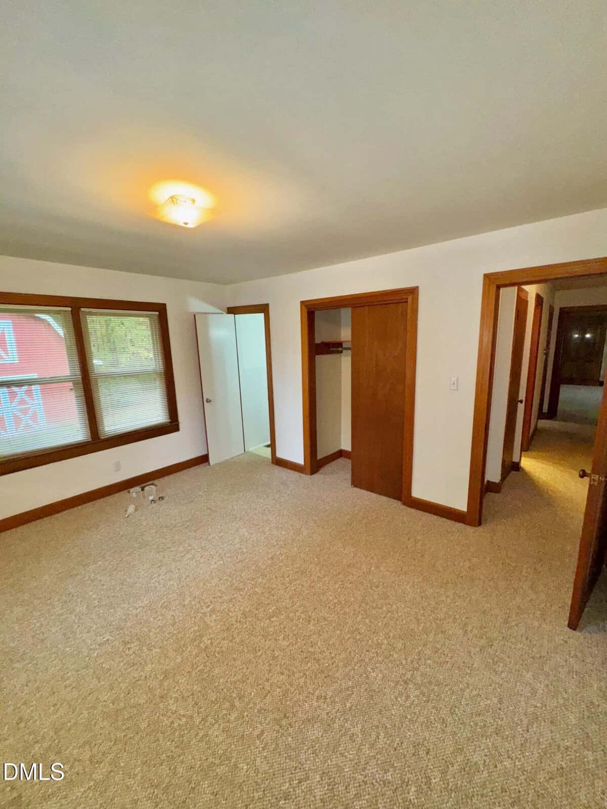 Empty carpeted bedroom with white walls, wood trim, a window, closet, open door leading to a hallway, and another door opening to a small room. A pair of shoes is visible on the floor near the window.