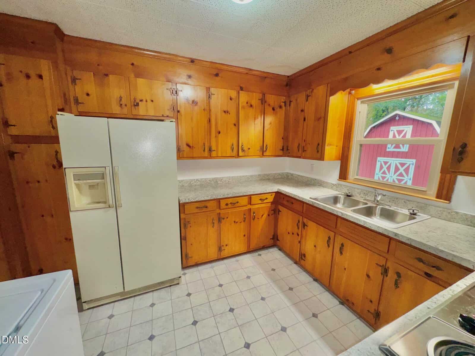 A kitchen with natural wood cabinets, a white refrigerator with ice dispenser, marble-patterned countertops, a double stainless steel sink under a window, and a red barn visible outside. The floor is white with a tile pattern.